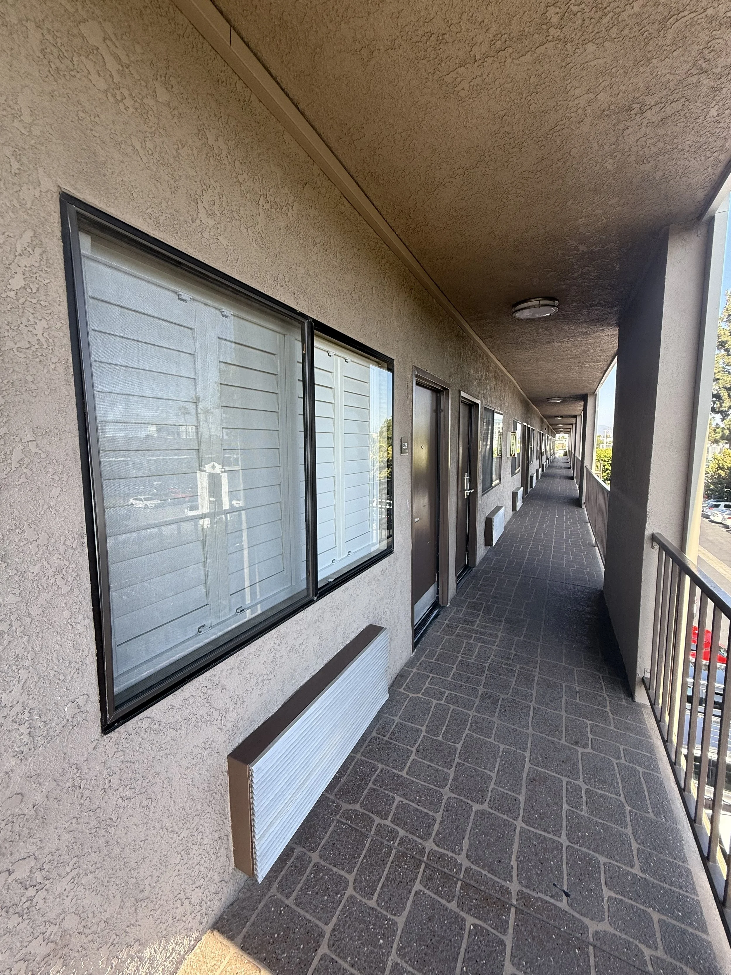 An outdoor corridor on a residential or apartment building with multiple doors and windows, white exterior walls, brick-patterned concrete flooring, and metal railings on the right.