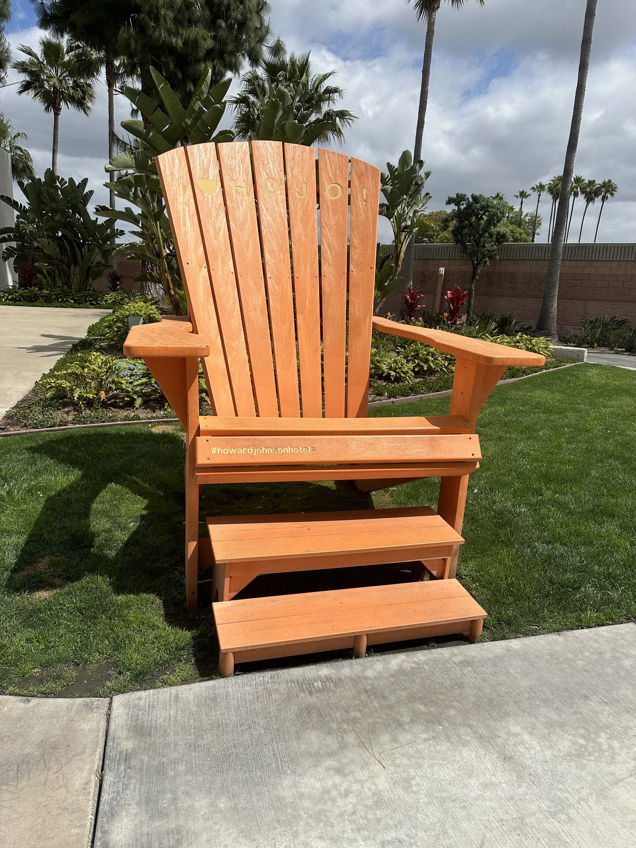 A large orange wooden Adirondack chair with steps in front of it, situated on a patch of green grass with palm trees and a sunny sky in the background.