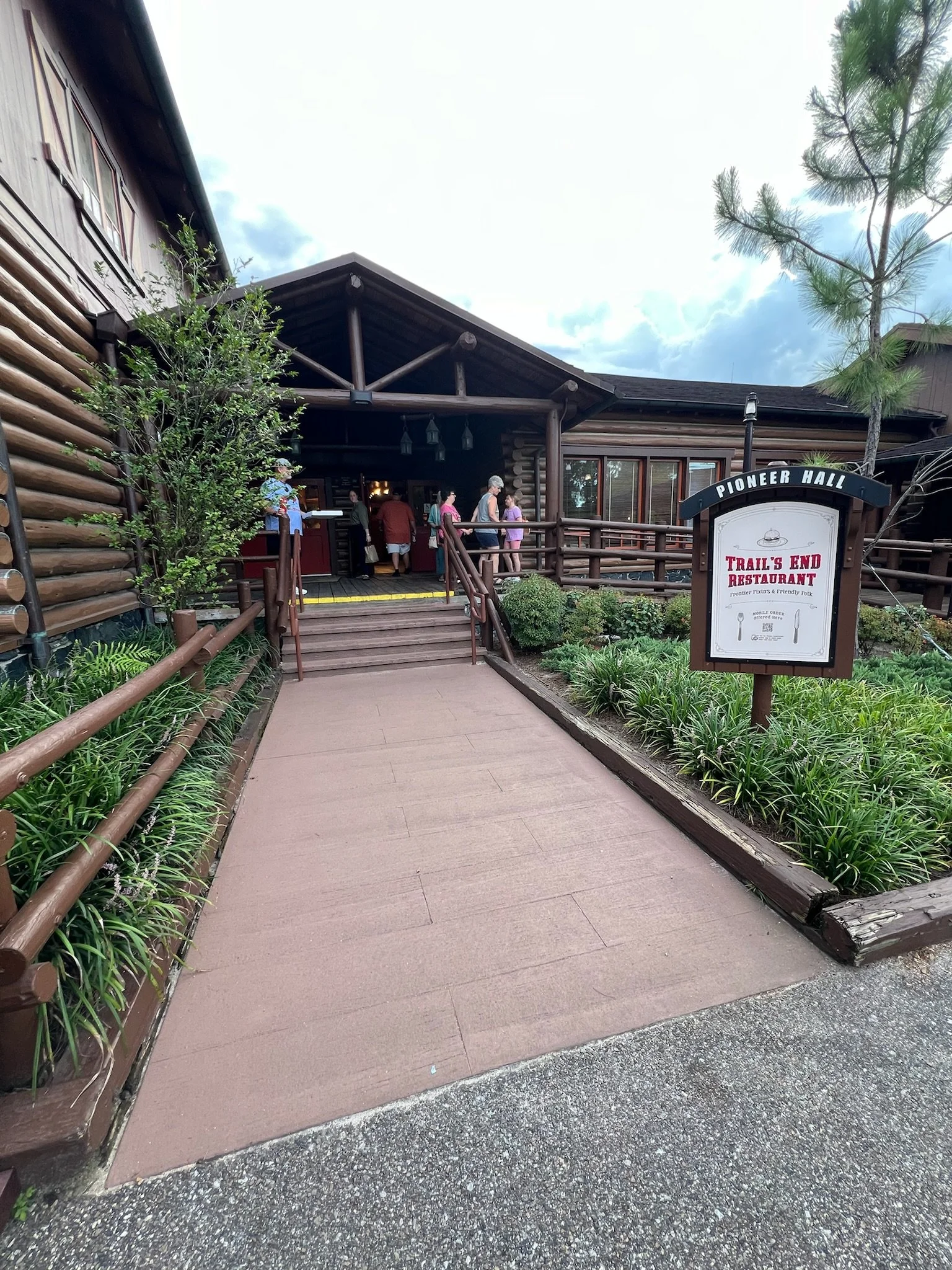 People entering the Trail's End Restaurant at Pioneer Hall, a rustic log building with a wooden deck, surrounded by landscaped greenery and a sign indicating the restaurant's name.