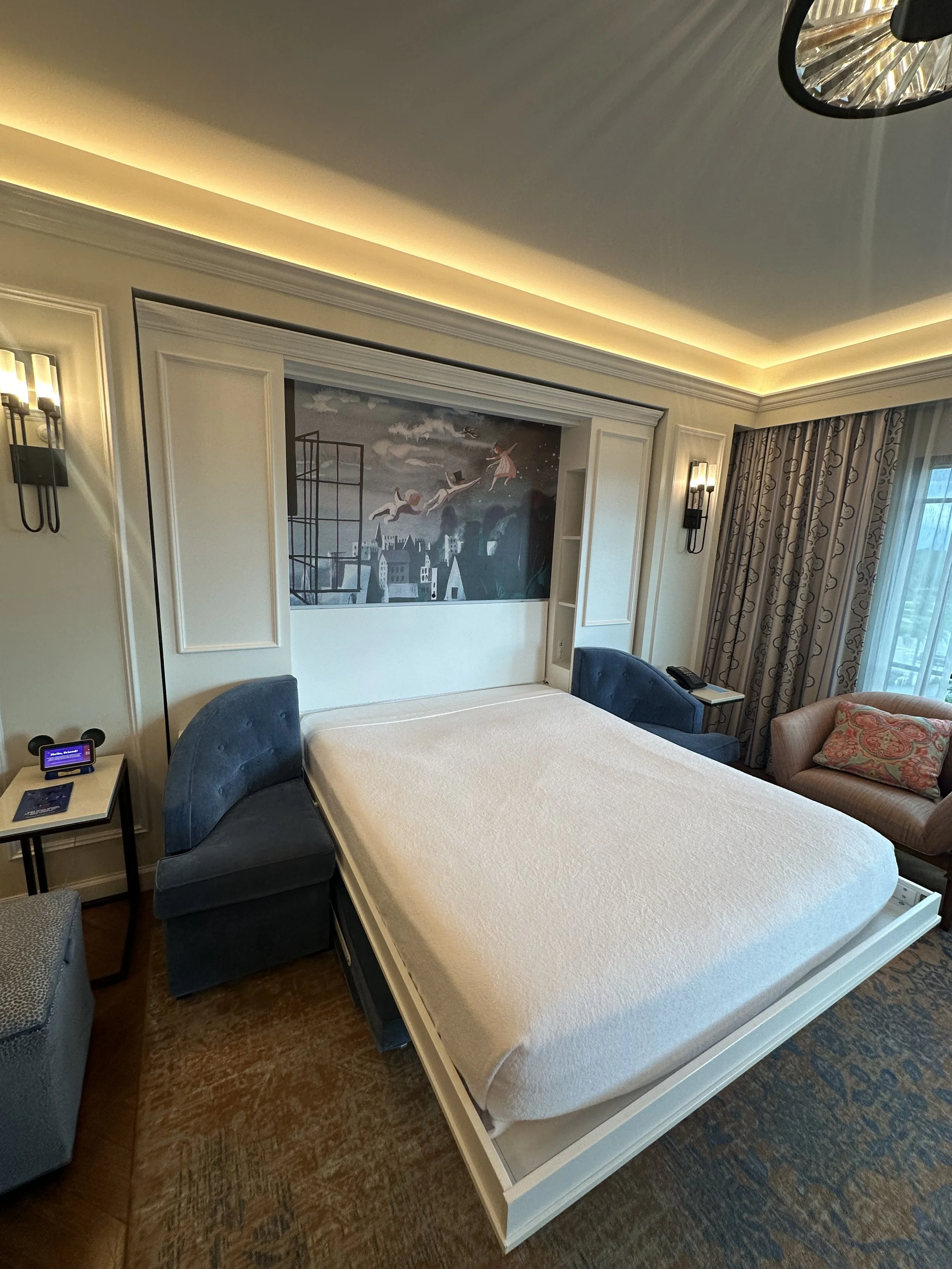 Hotel room with a large bed, two blue armchairs, a pink sofa with a patterned pillow, and artwork on the wall above the bed.