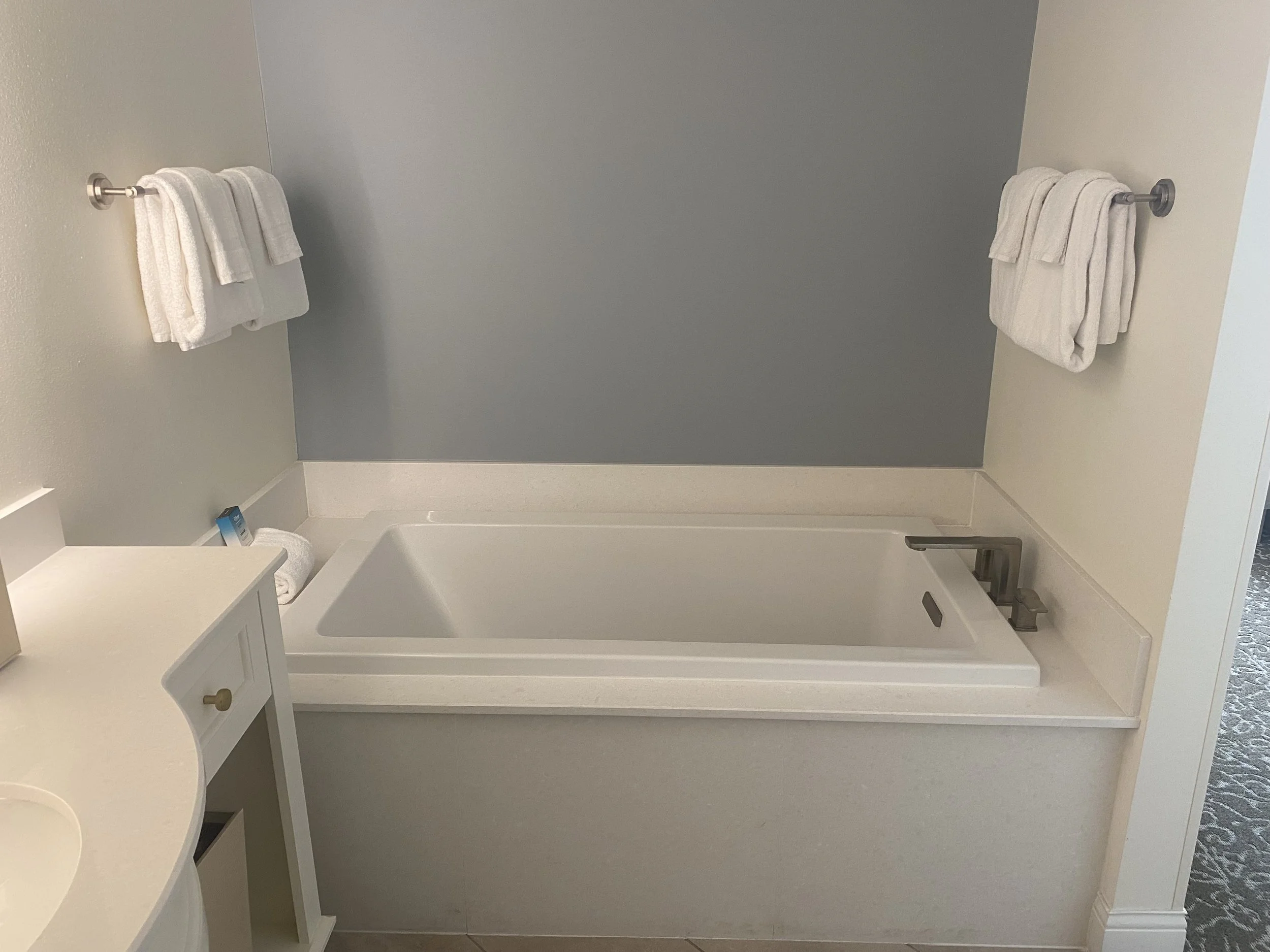 Bathroom with a white bathtub, two white towels on each side on towel bars, part of a white cabinet in the lower left, and a white wall on the right with a patterned carpet visible in the doorway.