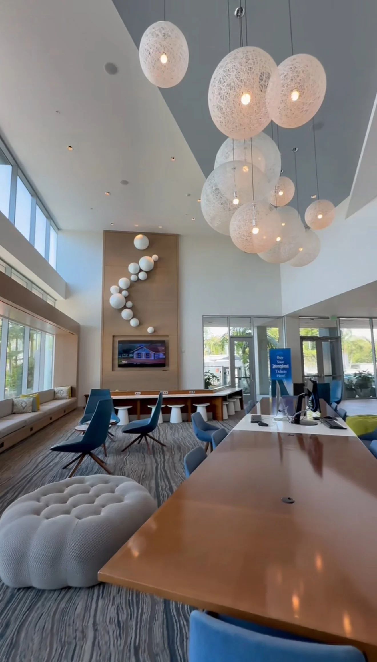 Hotel lobby with modern design featuring large spherical pendant lights, a reception desk with chairs, a wall with decorative white spheres, and large windows bringing in natural light.