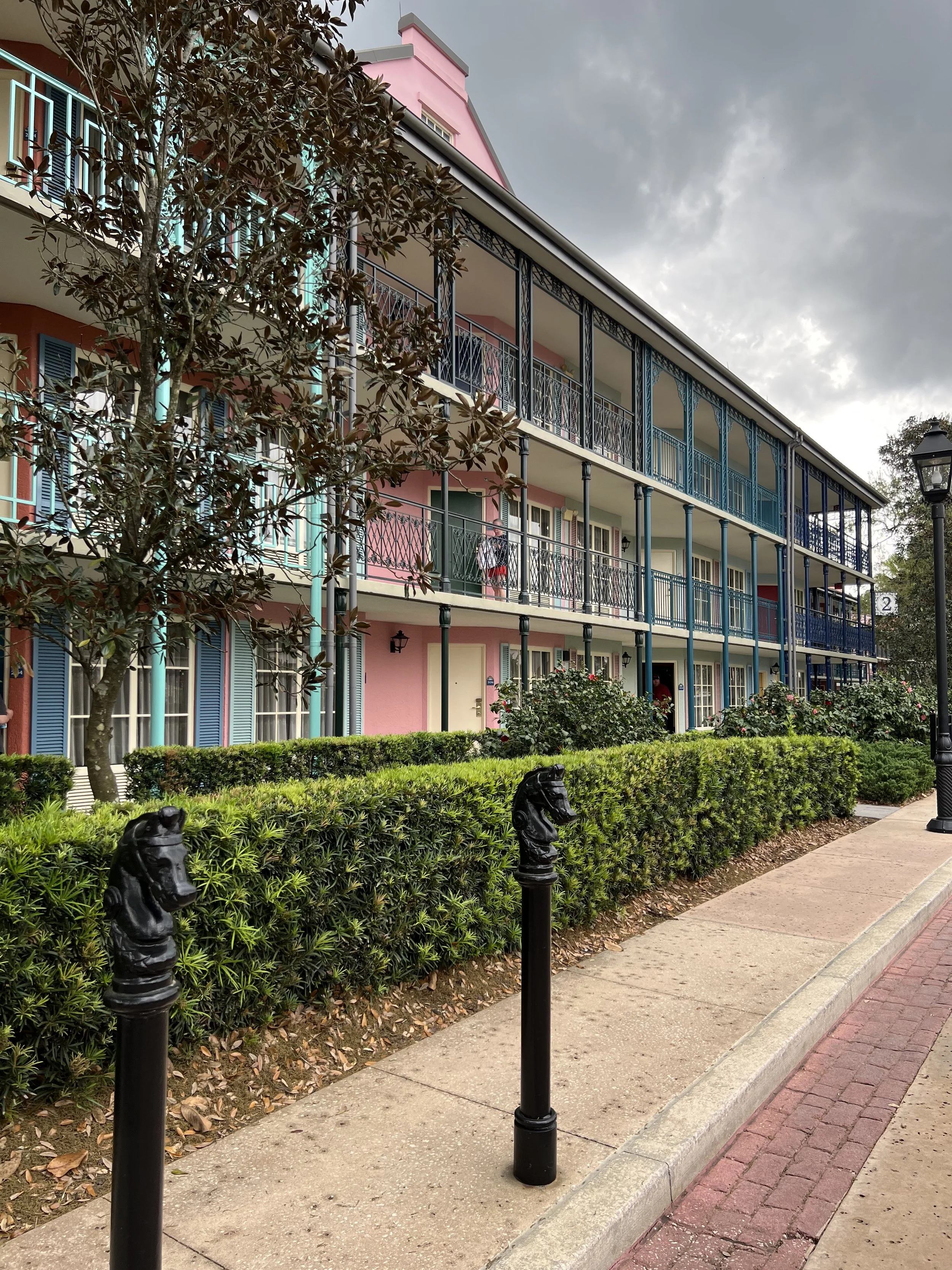 Colorful three-story apartment building with pink, blue, and green exterior, metal railings on balconies, surrounded by green bushes and a sidewalk with decorative black horse head posts, under a cloudy sky.