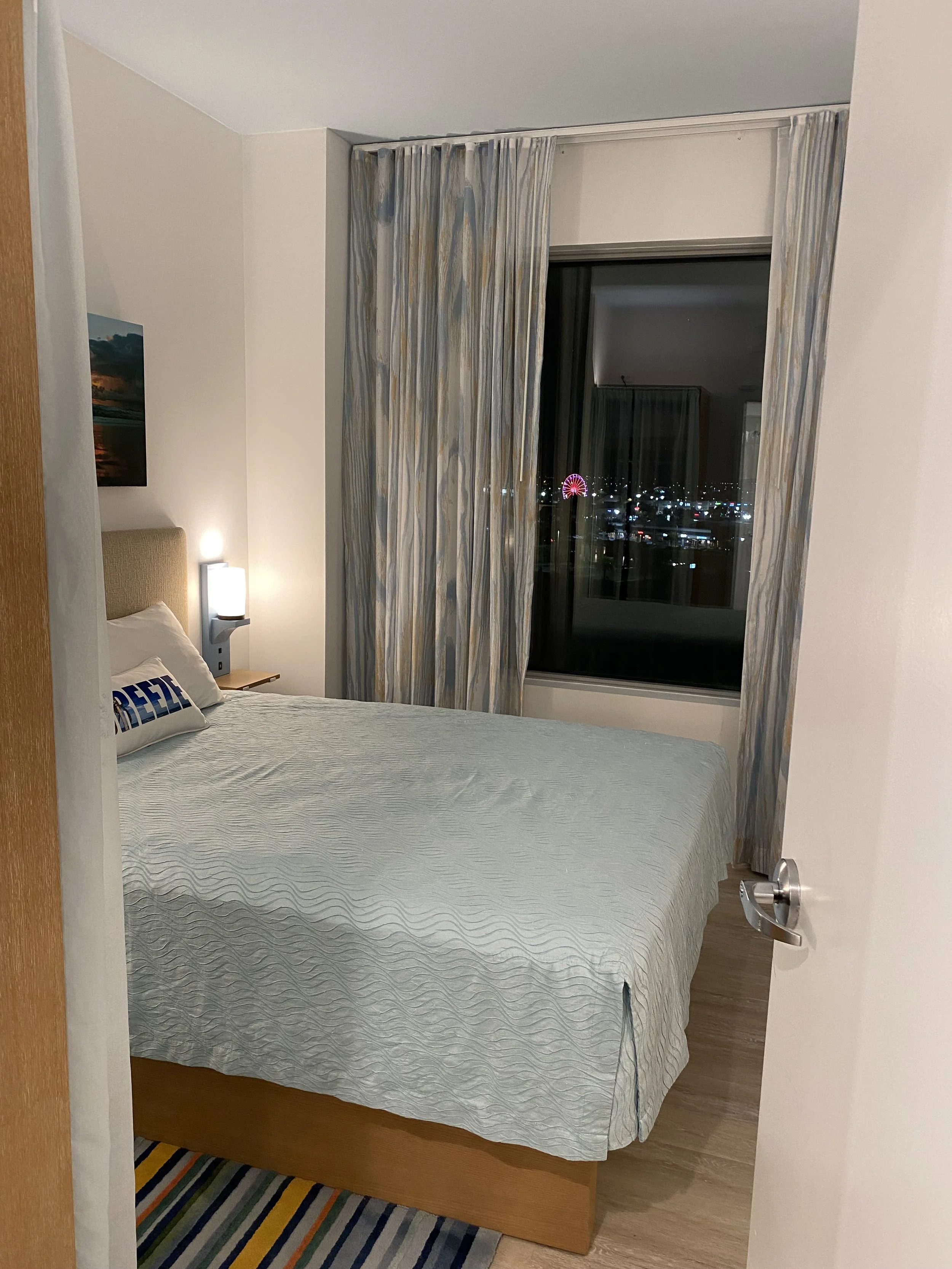 A hotel room with a bed, a nightstand with a lamp, a window with closed patterned curtains, and a night view outside with city lights and a red Ferris wheel.