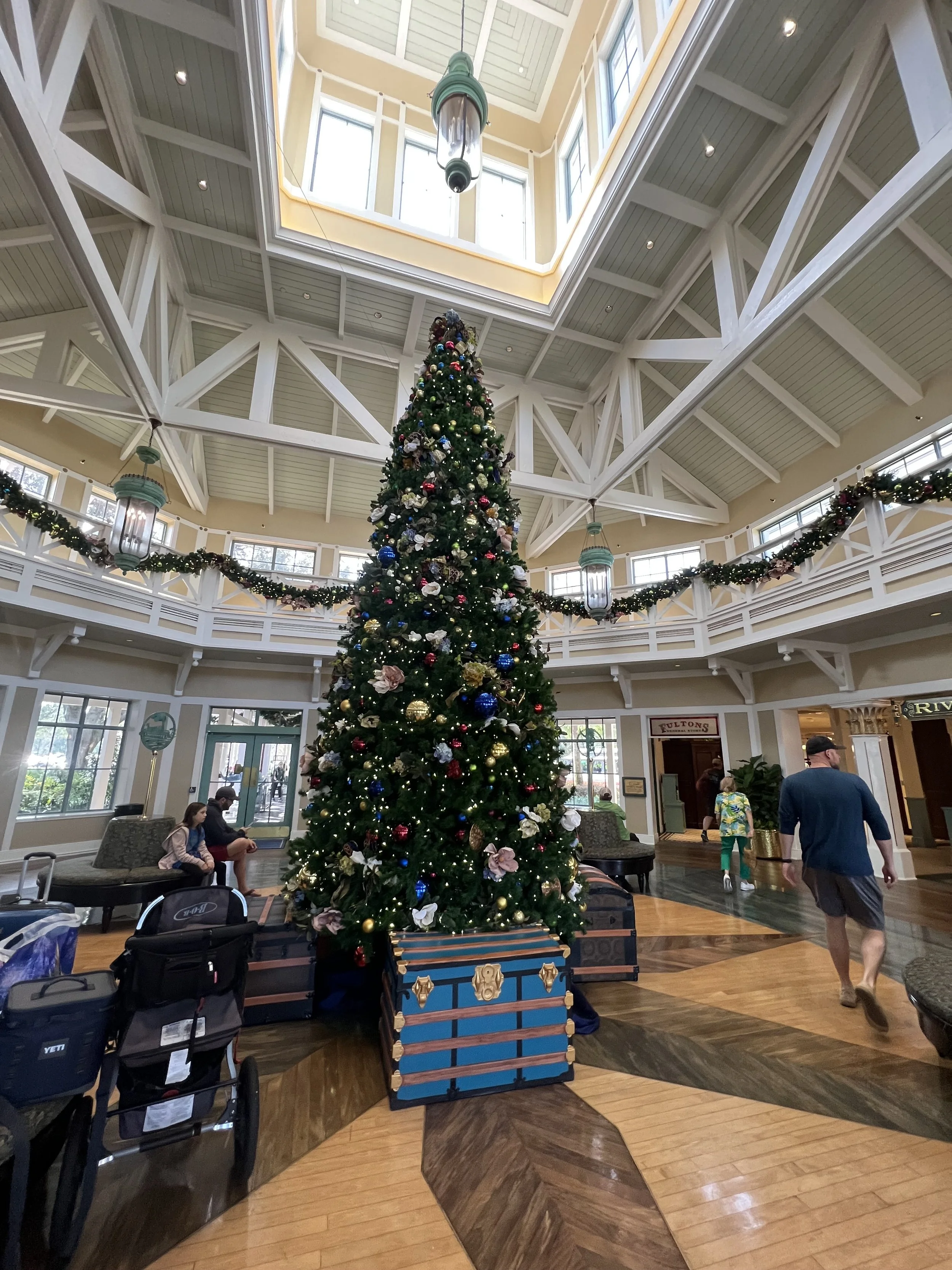 A large decorated Christmas tree in a spacious, well-lit hotel lobby with a high ceiling, white beams, and large windows. People are sitting and walking around, with holiday garlands and hanging lanterns.