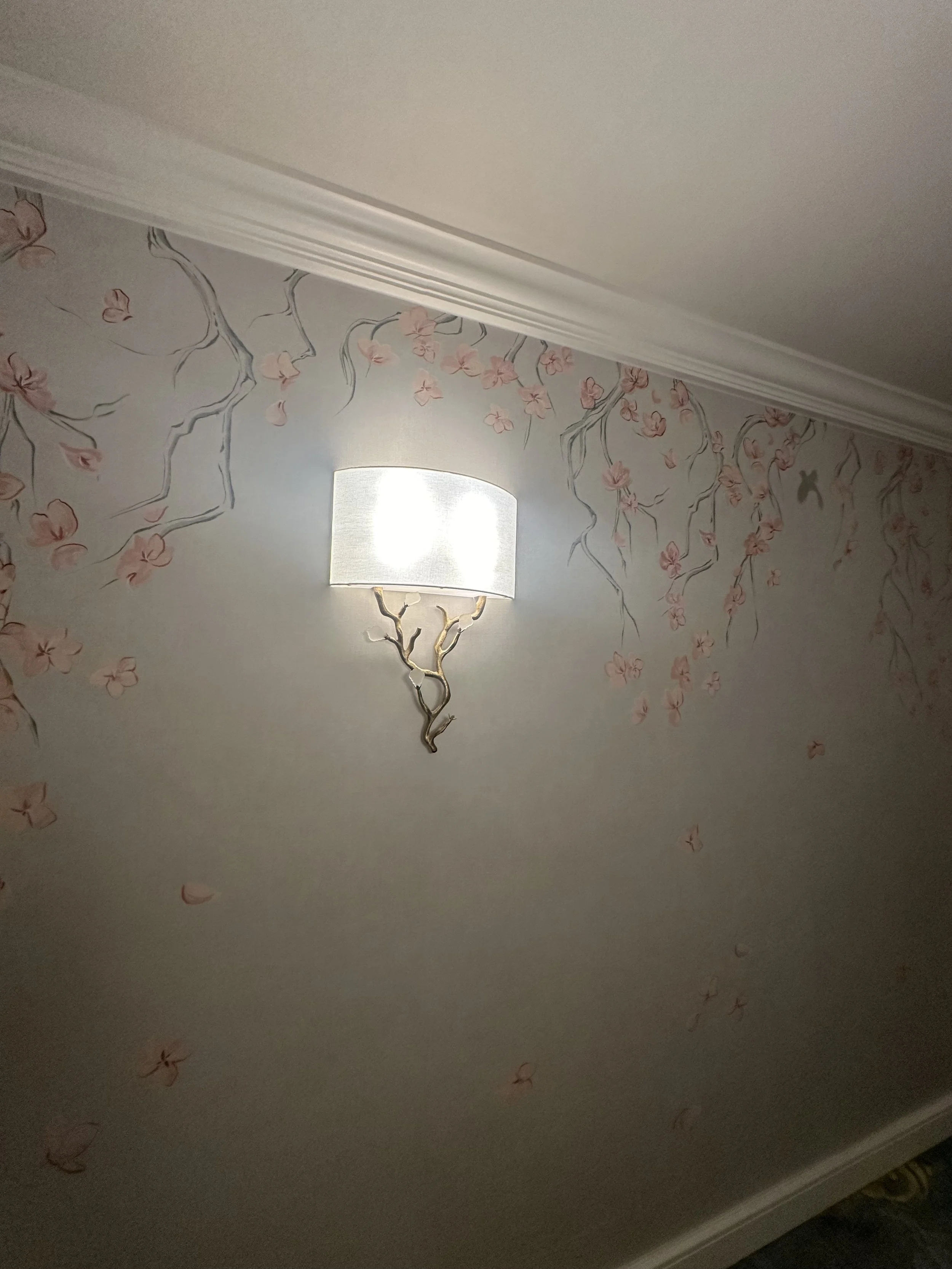Decorative wall with pink cherry blossom mural, white crown molding, and a wall-mounted lamp with a branch-shaped base.