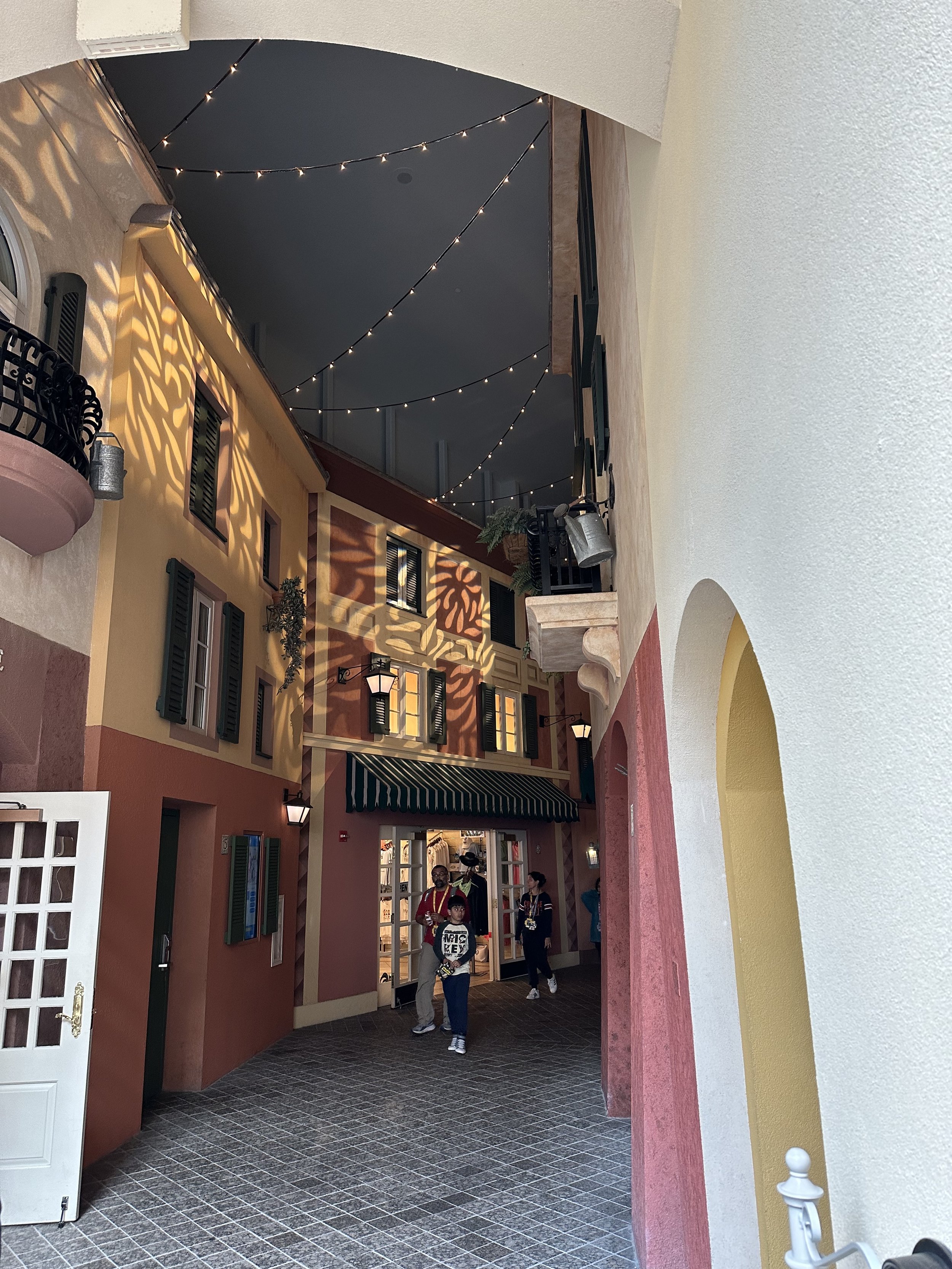 Indoor passageway with colorful buildings and string lights overhead, resembling a Mediterranean village.