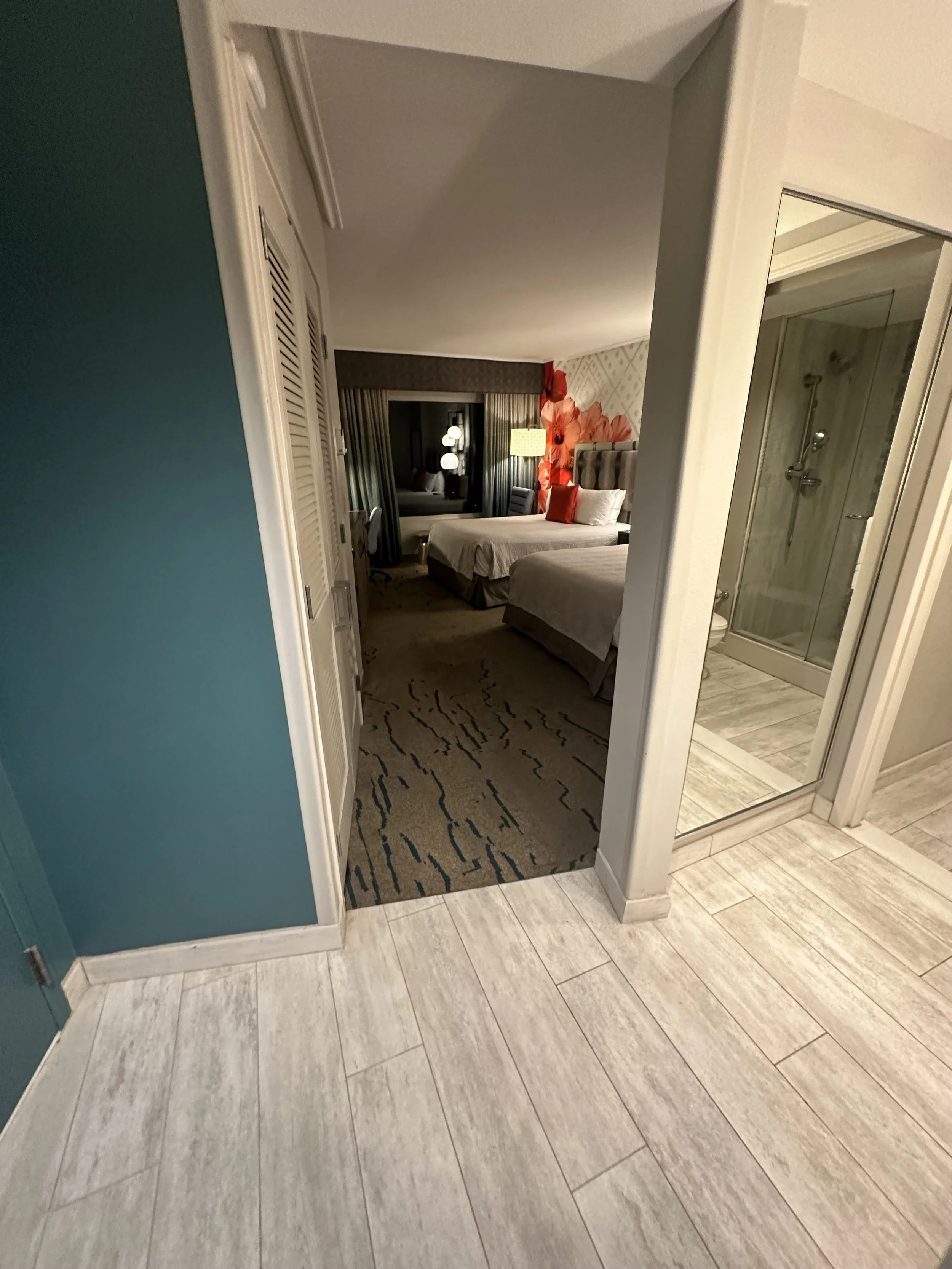 View from hallway into hotel room with two double beds, a window with curtains, a nightstand with a lamp, and a bathroom with a shower.