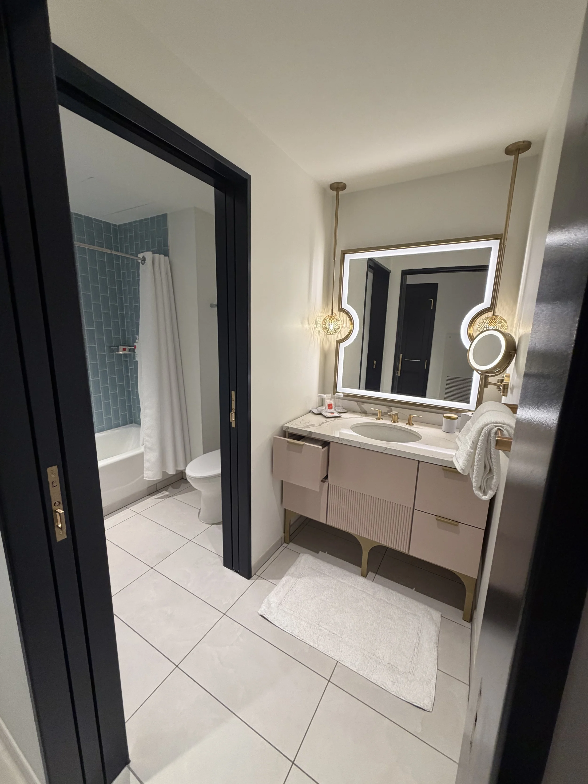 Modern bathroom with white walls, a vanity with a marble countertop, a backlit mirror, and gold accents. To the left, a shower area with blue subway tiles and a white shower curtain, separated by a black sliding door.