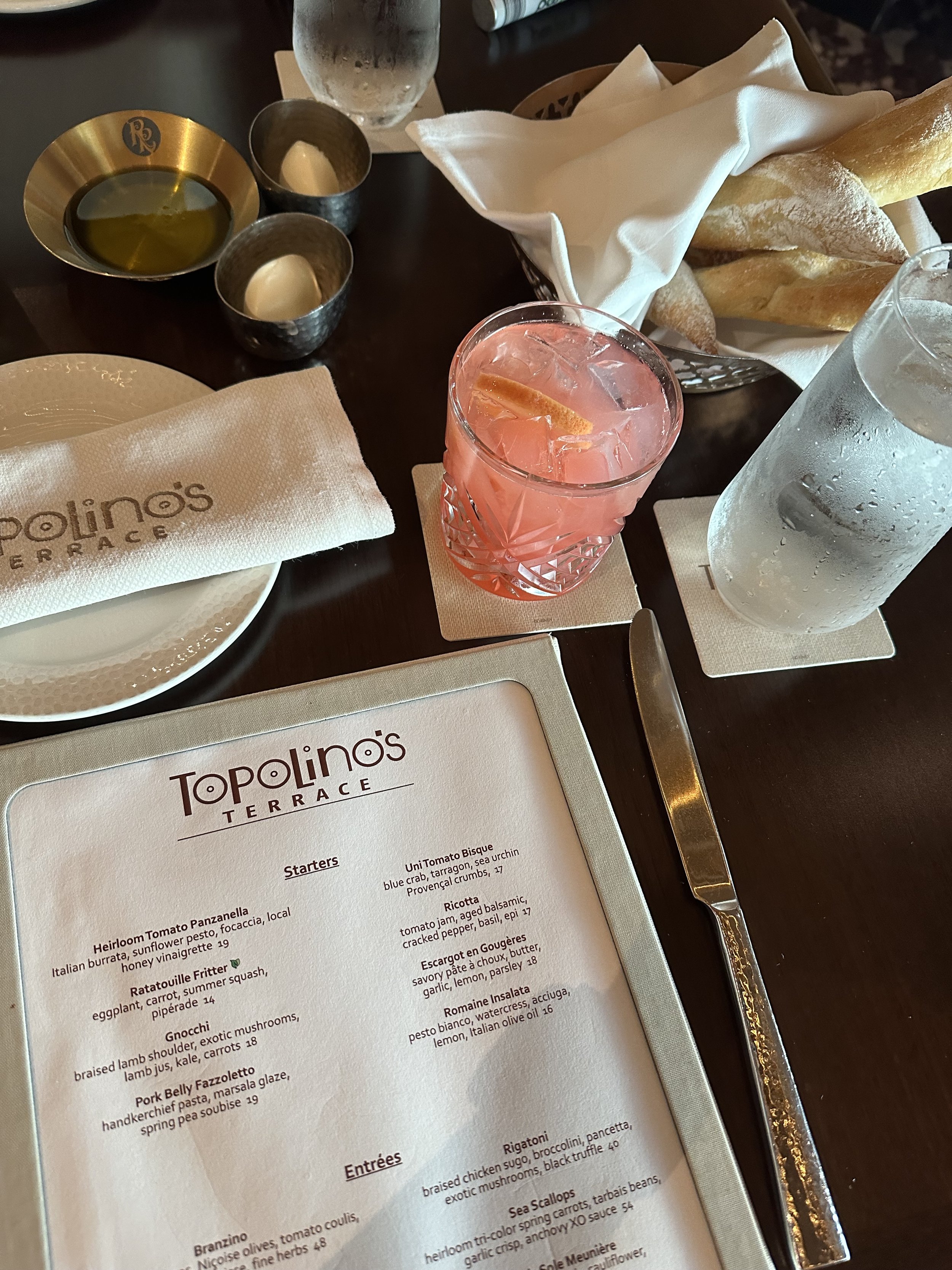 A restaurant table setting with Polino's Terrace menu, a pink cocktail with ice and a lemon wedge, a glass of water, a basket of breadsticks, small bowls with butter and olive oil, and a glass of water.