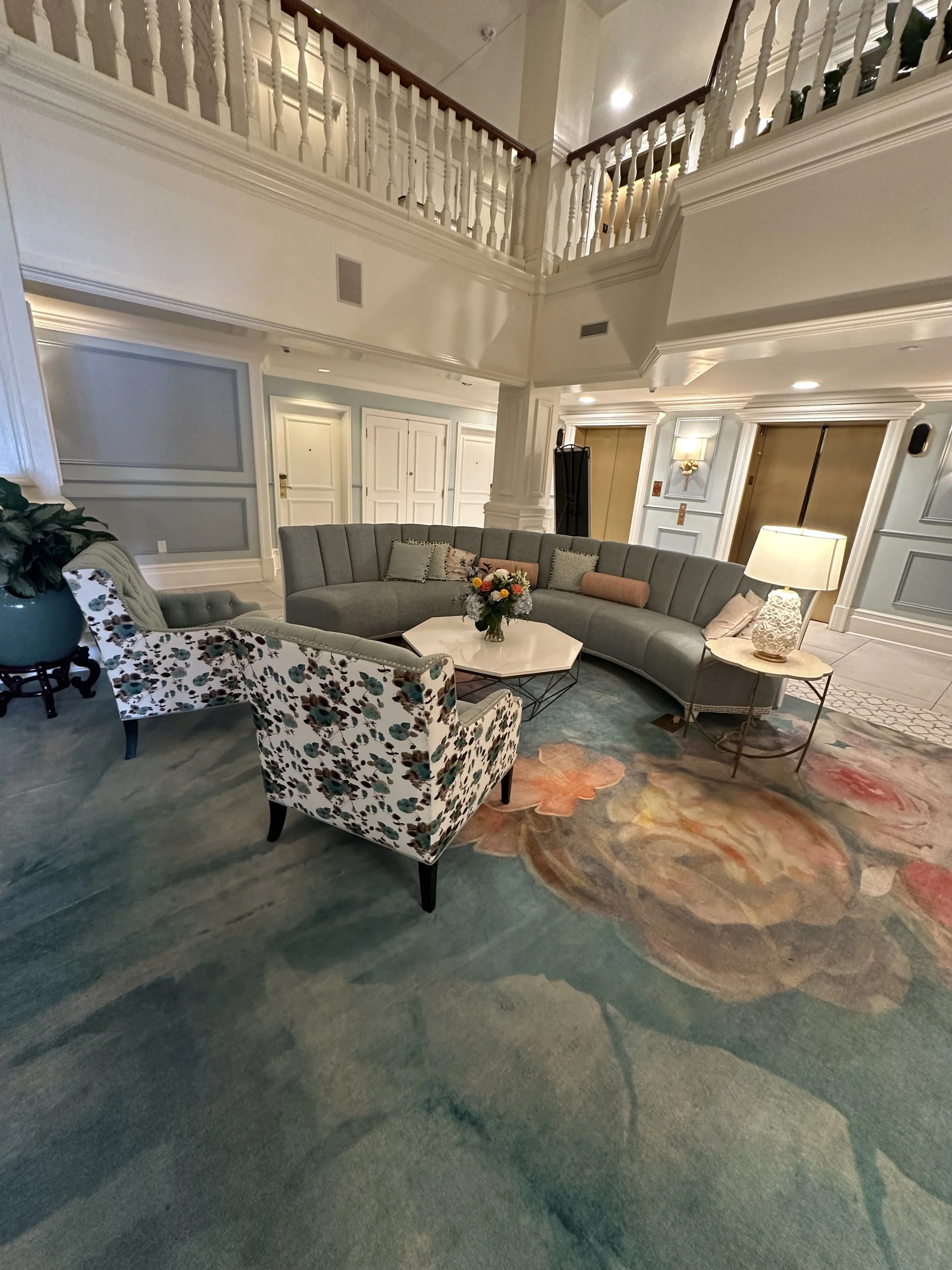 Luxury hotel lobby with floral carpet, curved gray sectional sofa, floral armchair, coffee table with flowers, side table with lamp, elevator doors, and upper balcony railing.