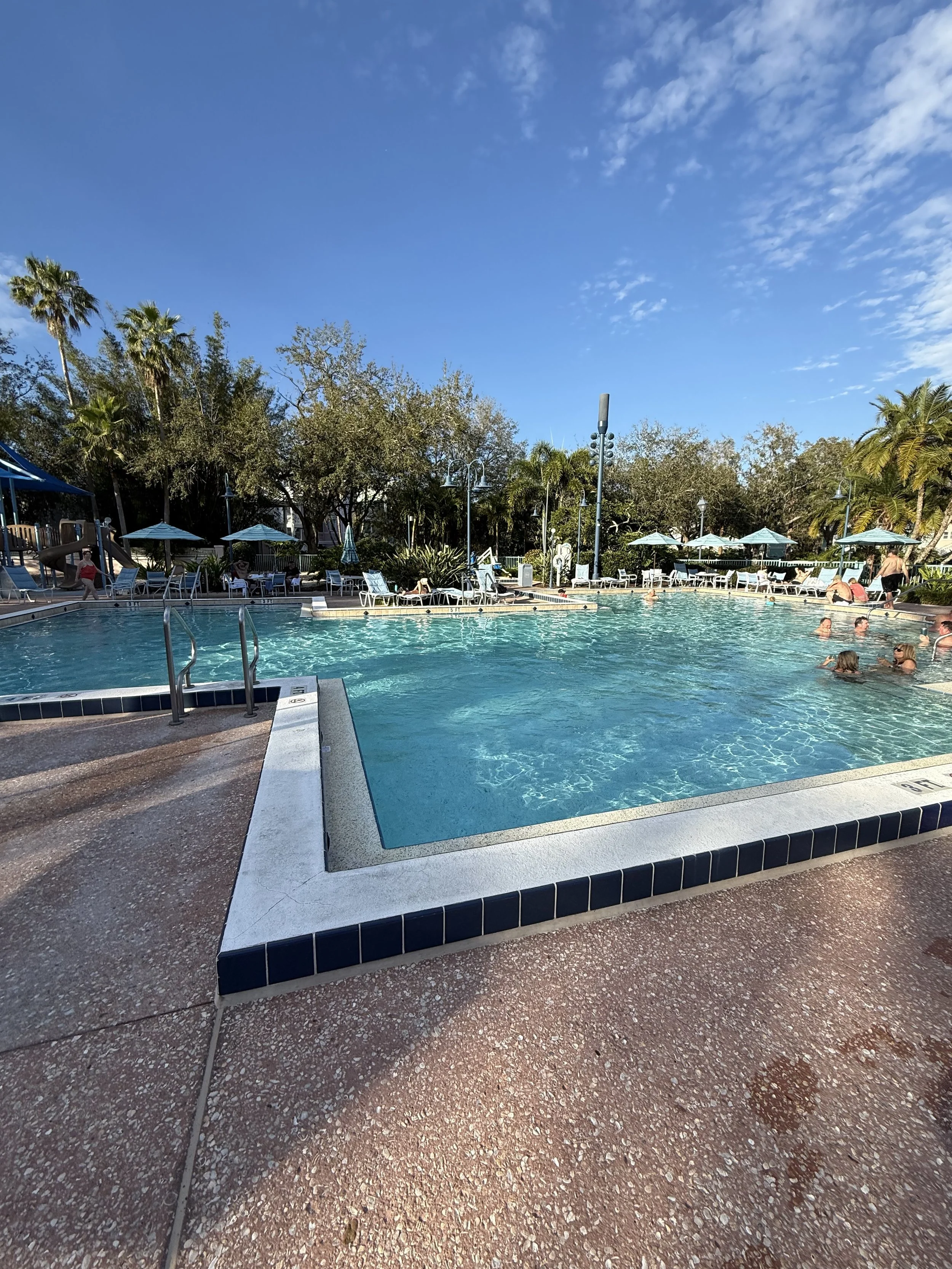 A swimming pool with people relaxing and swimming, surrounded by lounge chairs and green trees under a partly cloudy blue sky.