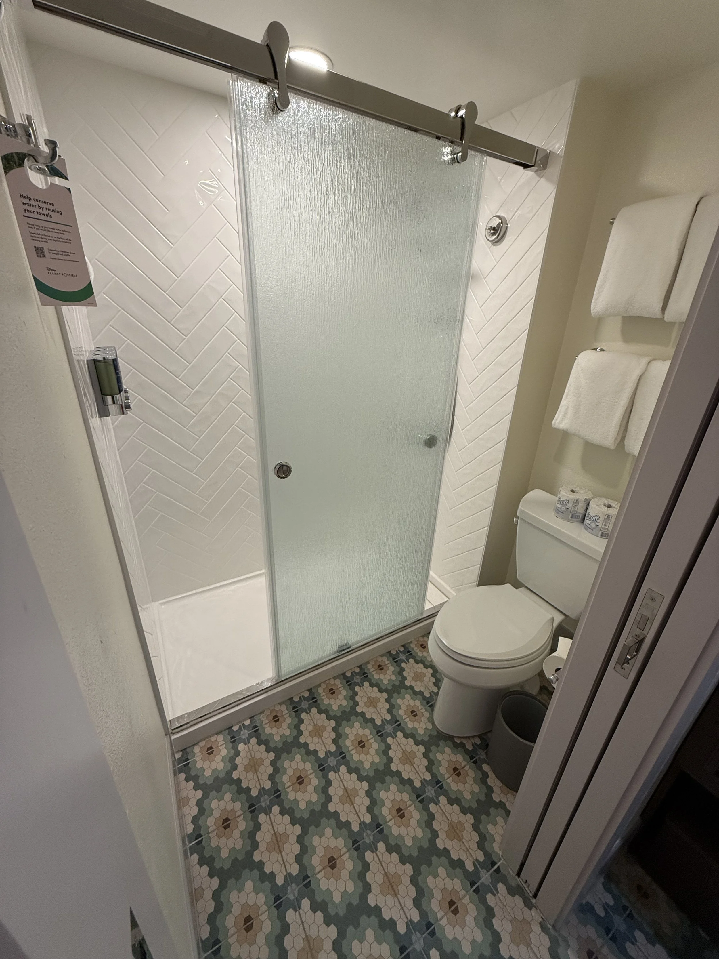 A small bathroom with a shower stall with frosted glass sliding door, a white toilet, and a colorful patterned tile floor. There are three white towels hanging on the wall and several toilet paper rolls on top of the toilet tank.