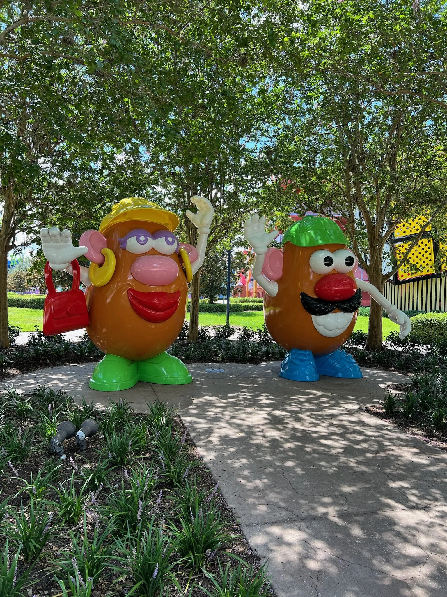 Two large, cartoon-like potato characters with exaggerated facial features, standing outdoors under a tree. One character has glasses, earrings, a yellow hat, green shoes, and a red bag, while the other has a green cap, mustache, and blue shoes. They