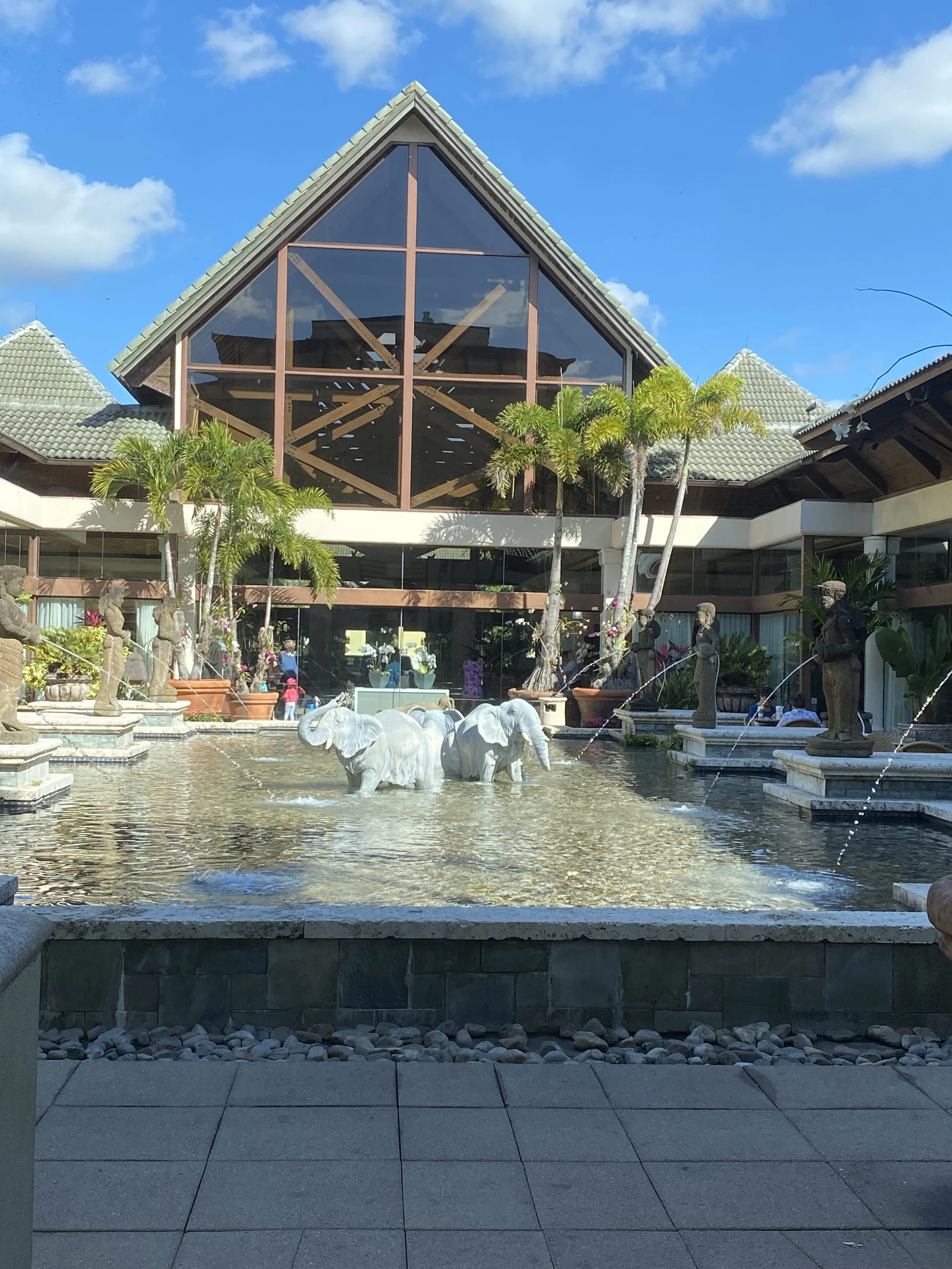 A building with a large triangular glass front, landscaped with palm trees, sculptures of elephants and people, and a water fountain with elephant statues in the foreground.