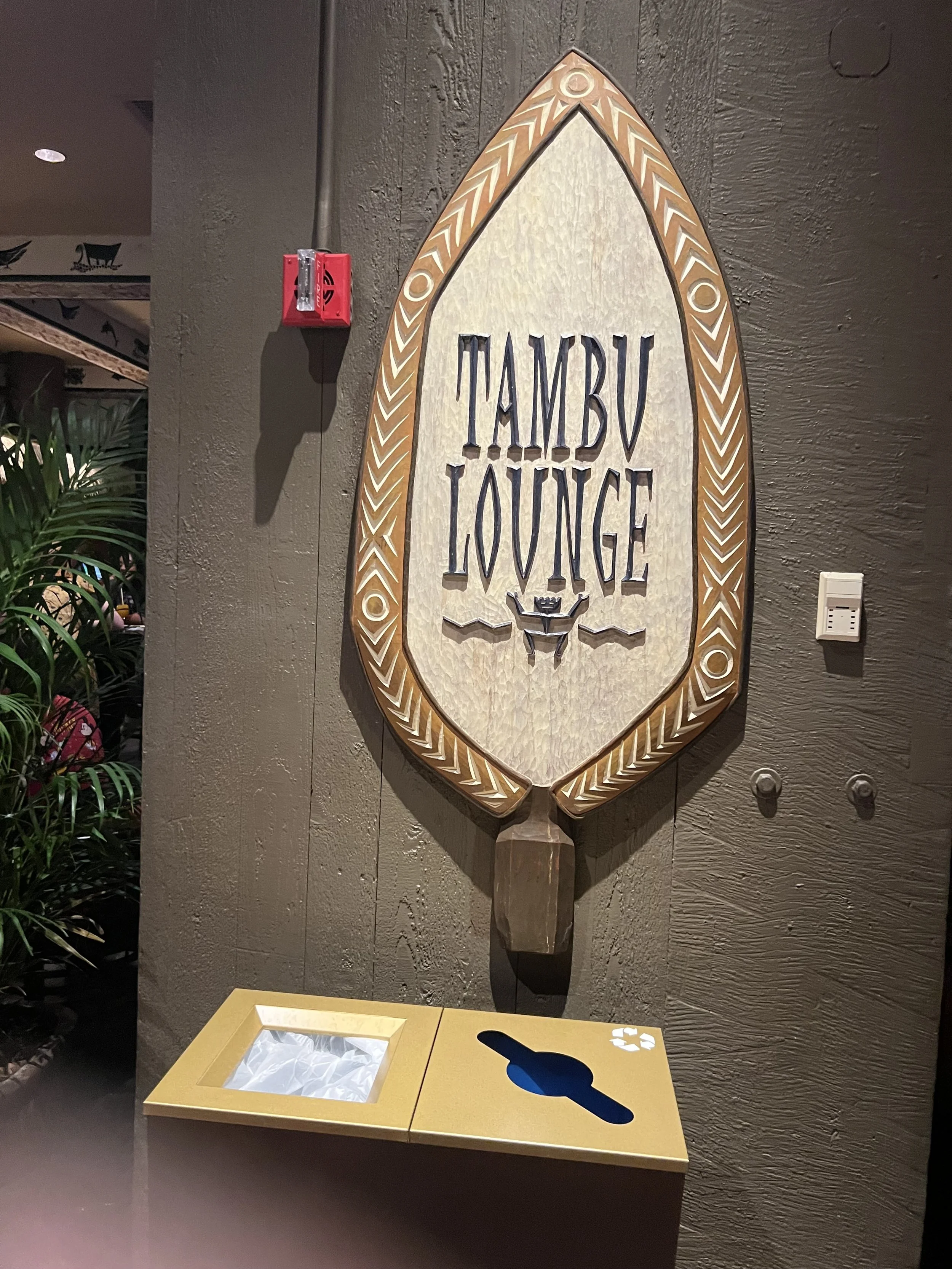 A large decorative sign for the 'TAMBU LOUNGE' mounted on a dark textured wall, with a wooden frame and carved details, and a trash bin with a slot in front of it.