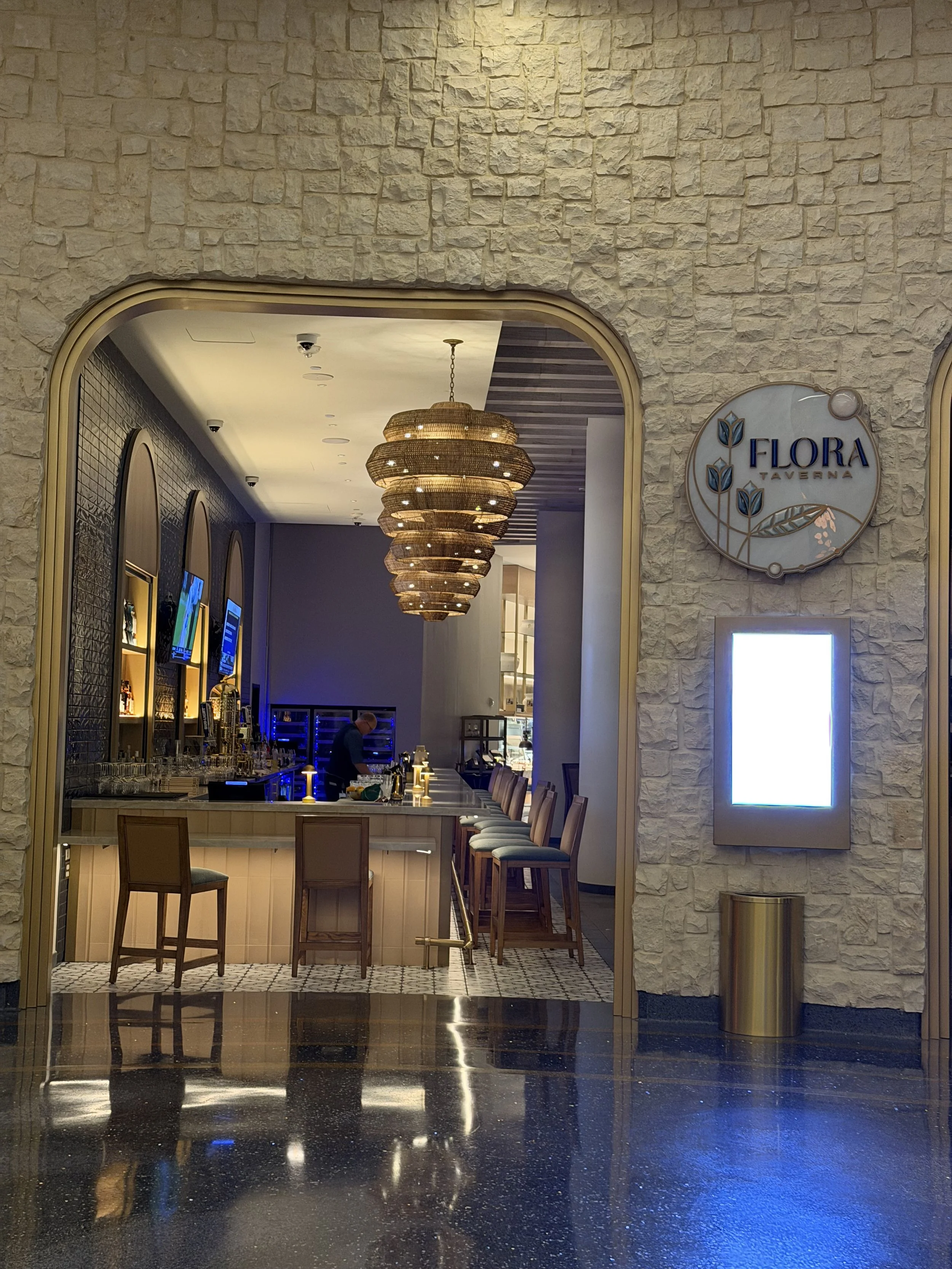 Interior view of Flora Taverna bar with a stone wall entrance, chandelier lighting, and a bartender behind the counter.