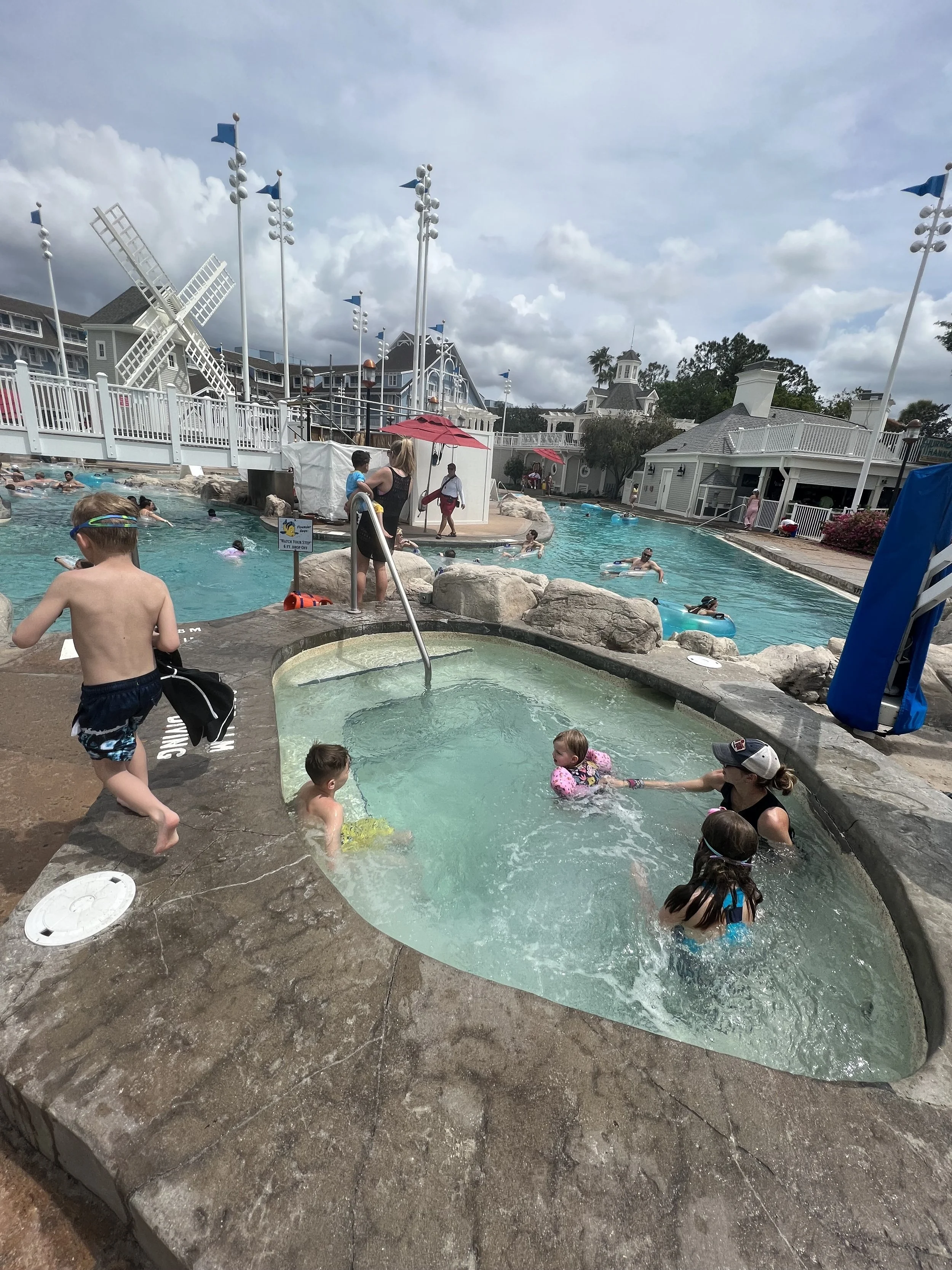 Children playing in a small pool with an adult supervising, while others swim and relax in a larger pool at a waterpark under a partly cloudy sky.