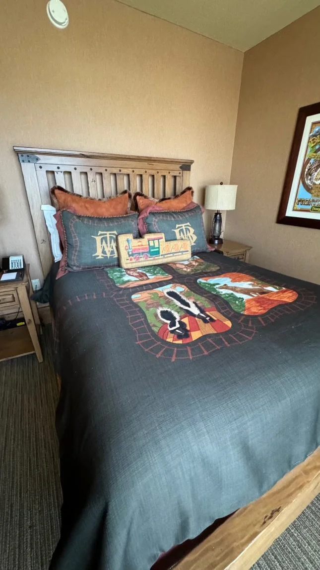 A hotel room with a wooden bed frame, two pillows with embroidered 'WD' initials, a decorative pillow shaped like a toy train, and a quilt featuring animals and nature scenes. There are two nightstands with lamps and a framed poster on the wall.
