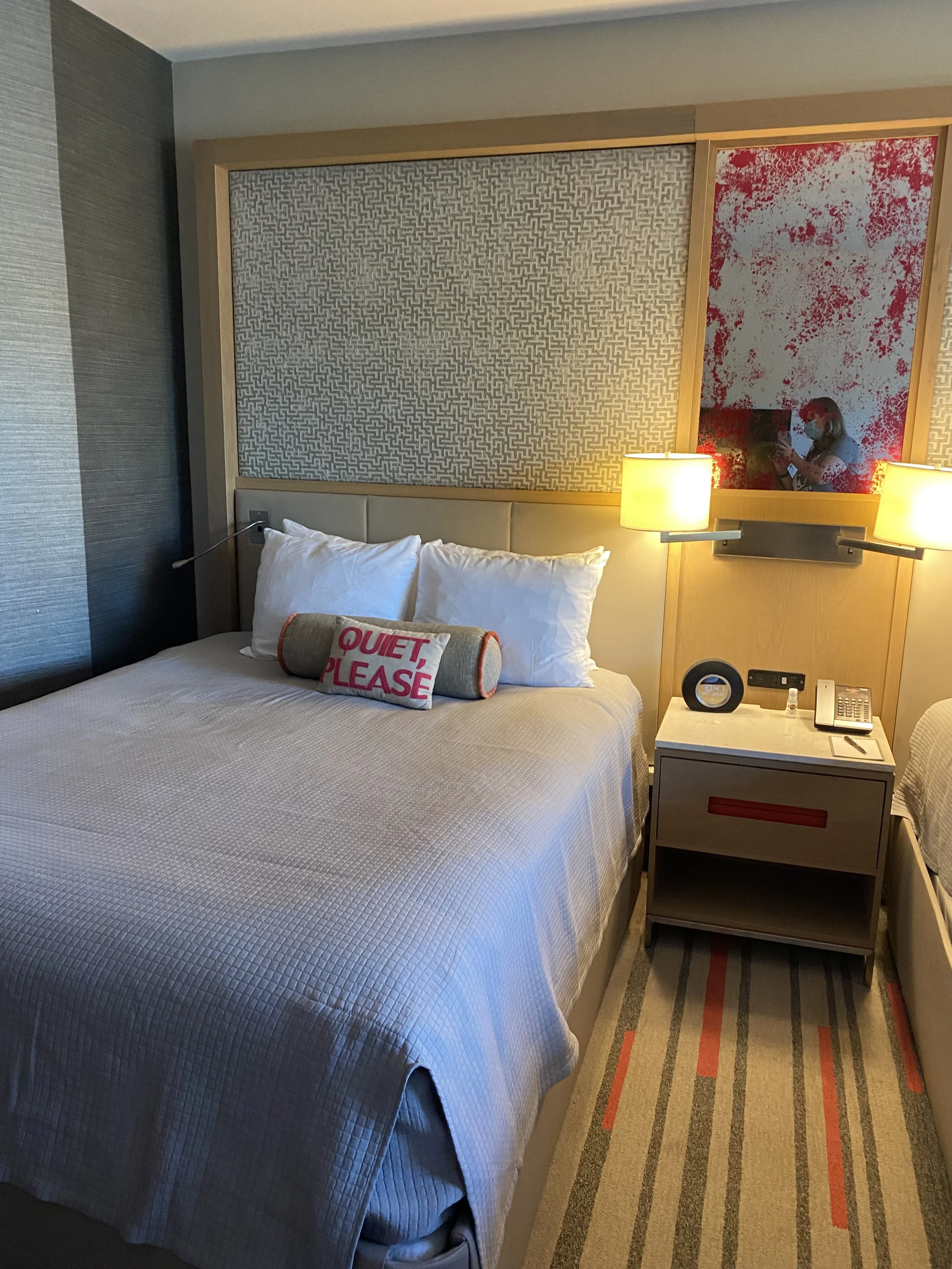 A hotel room with a bed featuring a pillow with the words "Quiet Please" embroidered on it, bedside lamps, a nightstand with a clock and phone, a large mirror with a person taking a photo, and patterned wall decor.