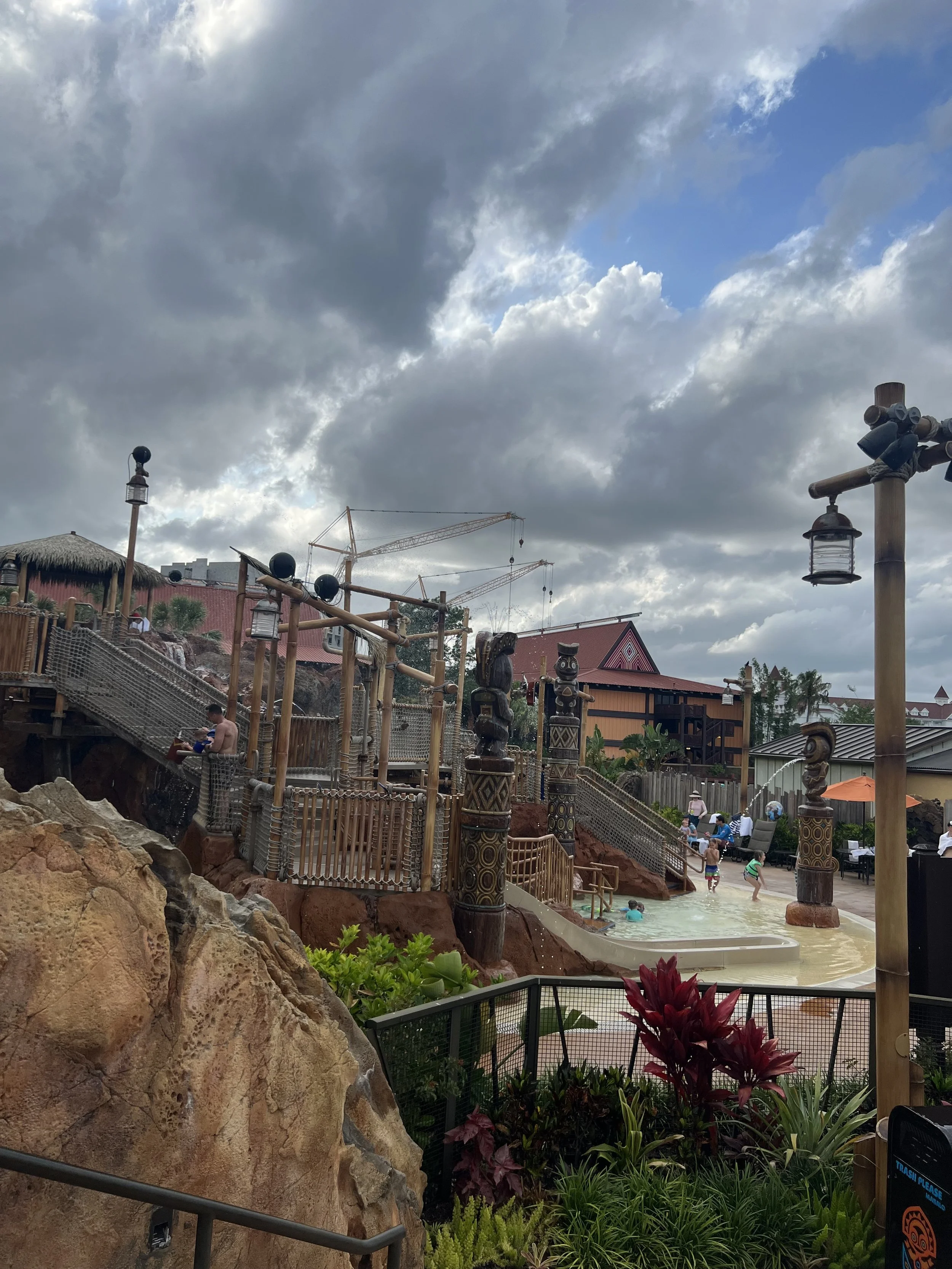 Water park with a rocky and wooden structure, children playing in a shallow pool, tropical plants, and cloudy sky.