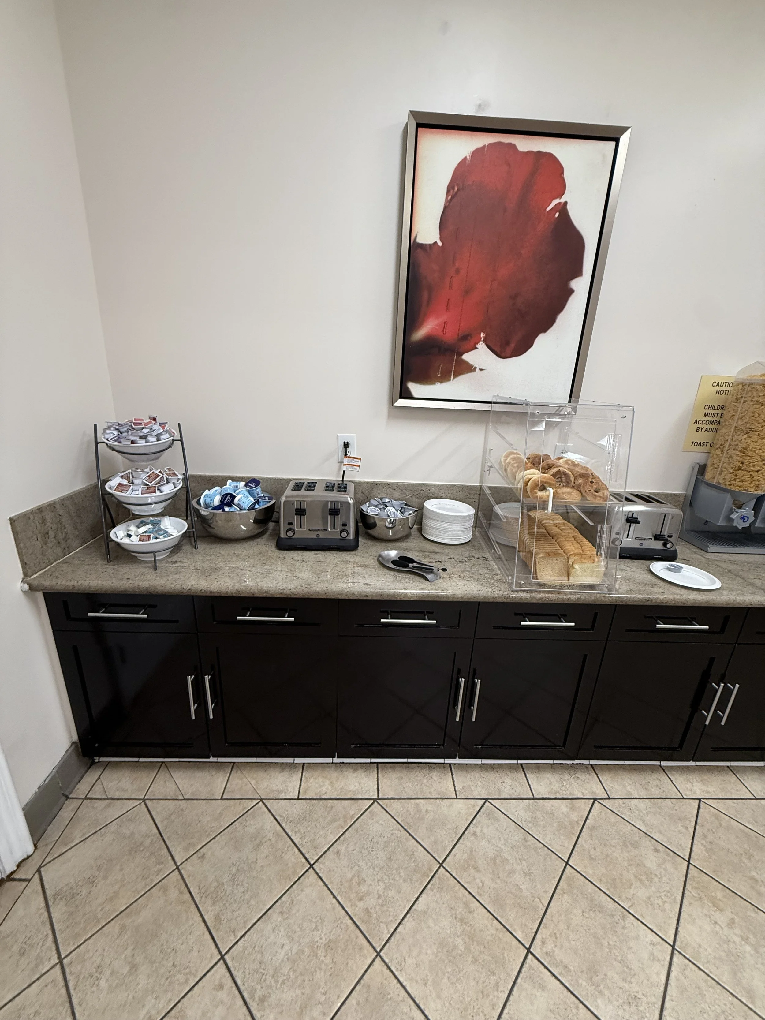 Snack bar area with a granite countertop, black cabinets, and a tiled floor. The counter has some condiments, a toaster, a stack of white plates, a container with pastries, a cereal dispenser, and a box of cereal. There is a piece of abstract art on 