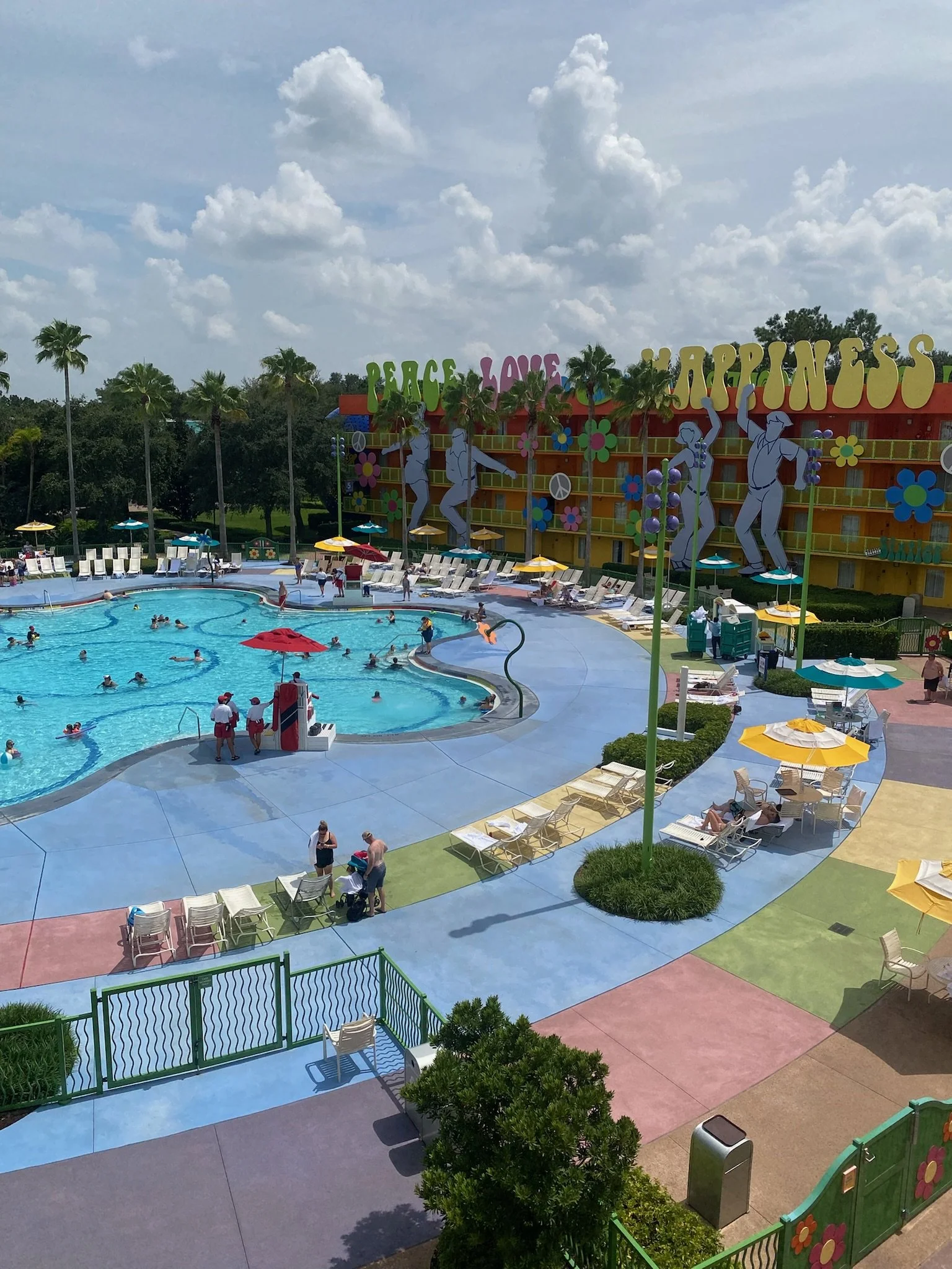An outdoor swimming pool area at a resort with children and adults swimming, lounging, and walking. The background features a colorful building with large signs saying 'PEACE LOVE HAPPINESS,' decorated with flower and peace sign motifs, surrounded by