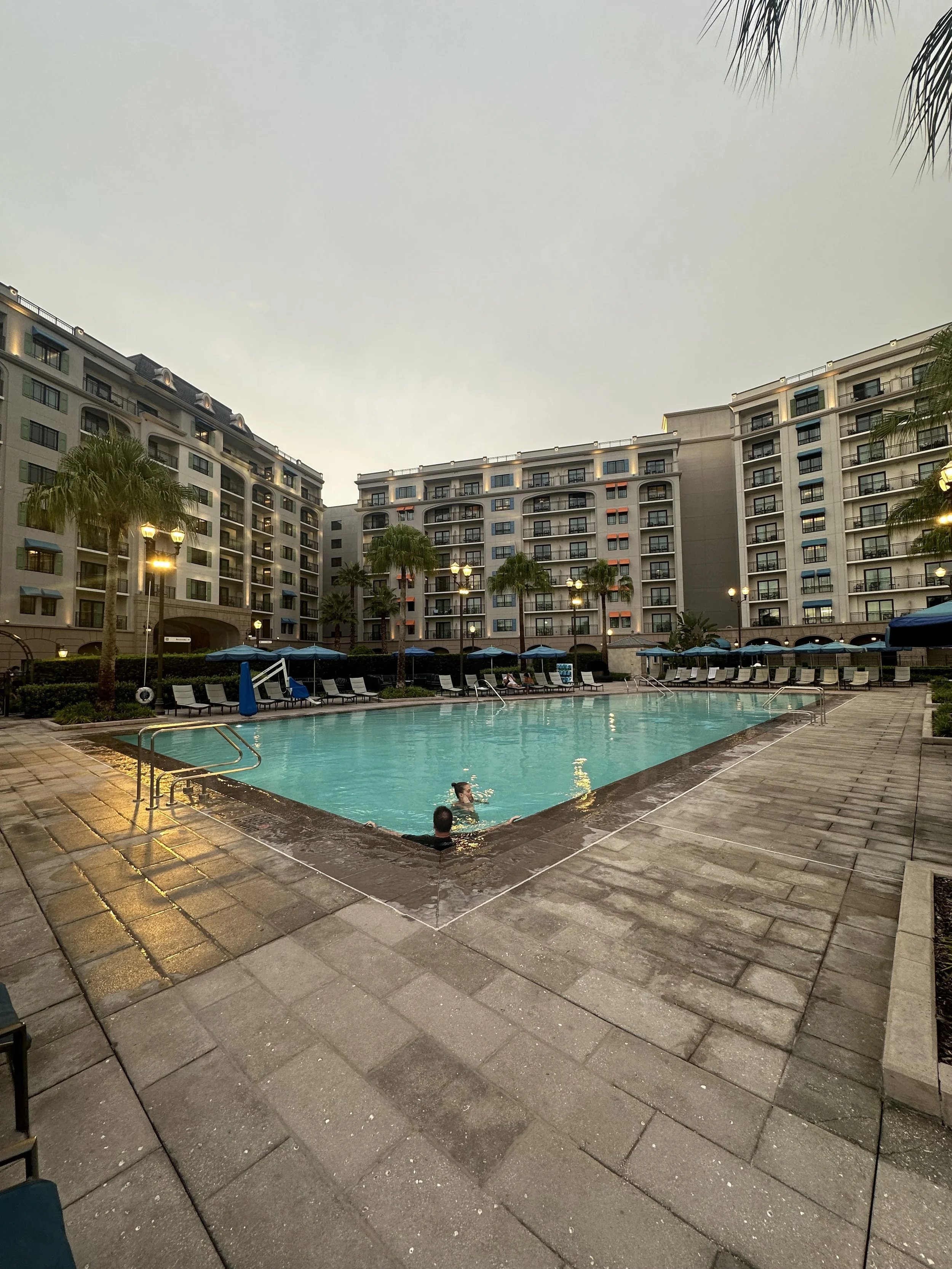 An outdoor swimming pool in a residential complex with a man and a child swimming. Poolside chairs and umbrellas line the deck, surrounded by palm trees and tall apartment buildings.