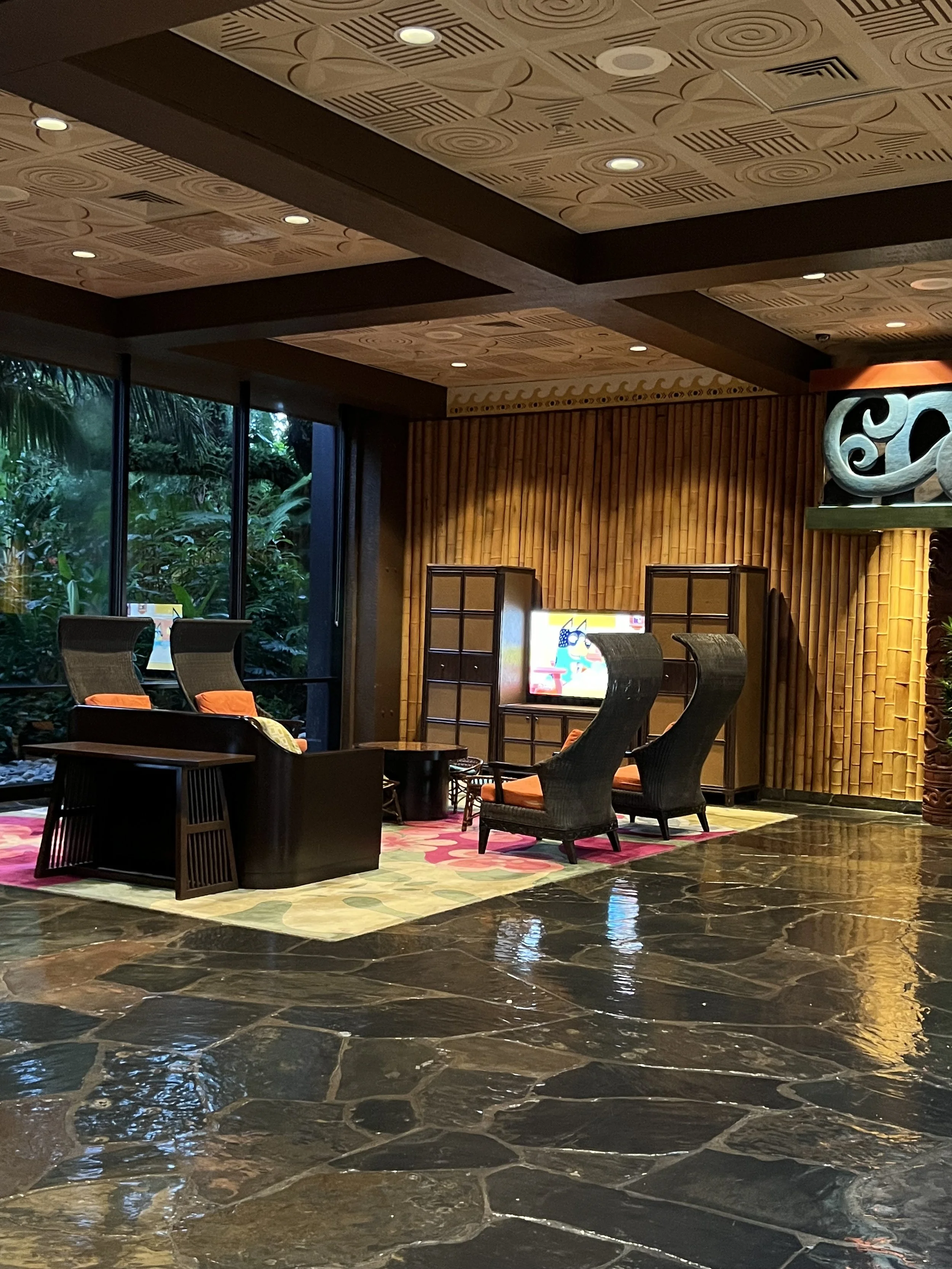 Indoor lounge area with wicker chairs, a small table, and a dresser. Large windows reveal lush green jungle outside. Wooden wall decor and a colorful rug complete the cozy, tropical ambiance.