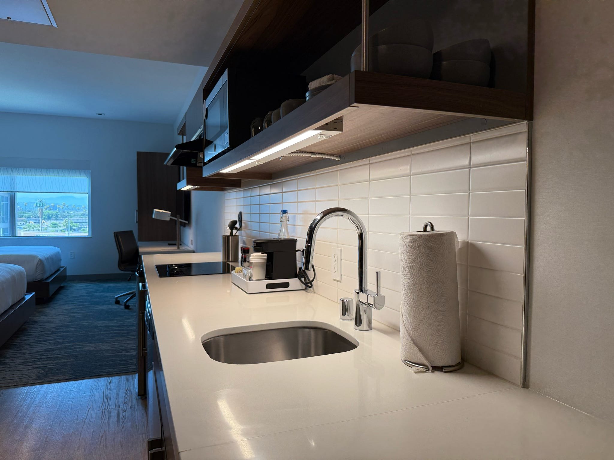 Modern kitchenette with a white countertop, stainless steel sink, faucet, and paper towel roll. White tile backsplash, dark wood upper cabinets, and view of a hotel room with beds and a window showing an outdoor cityscape.