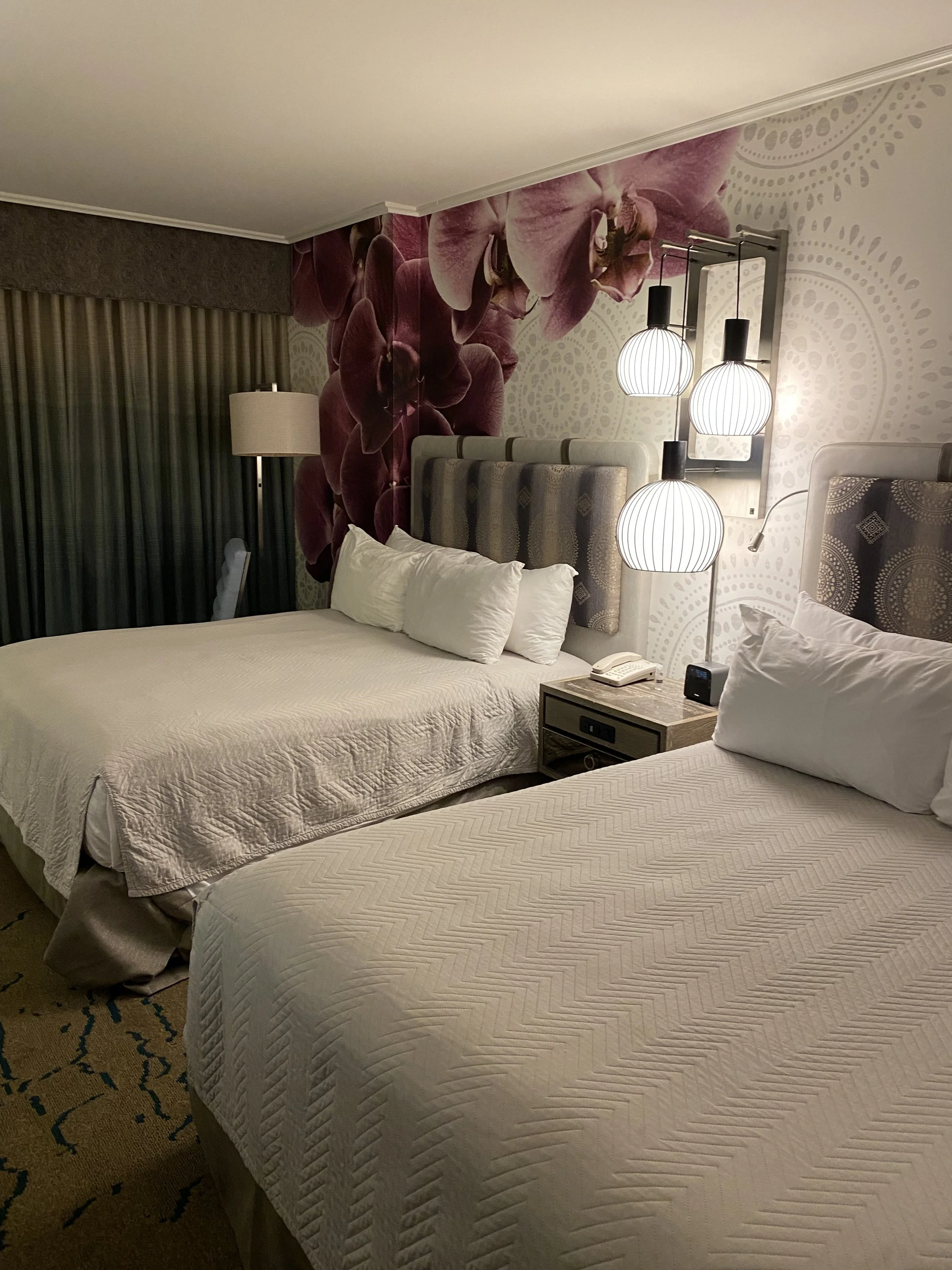 Hotel room with two beds, floral wall, nightstand, and hanging lamps.