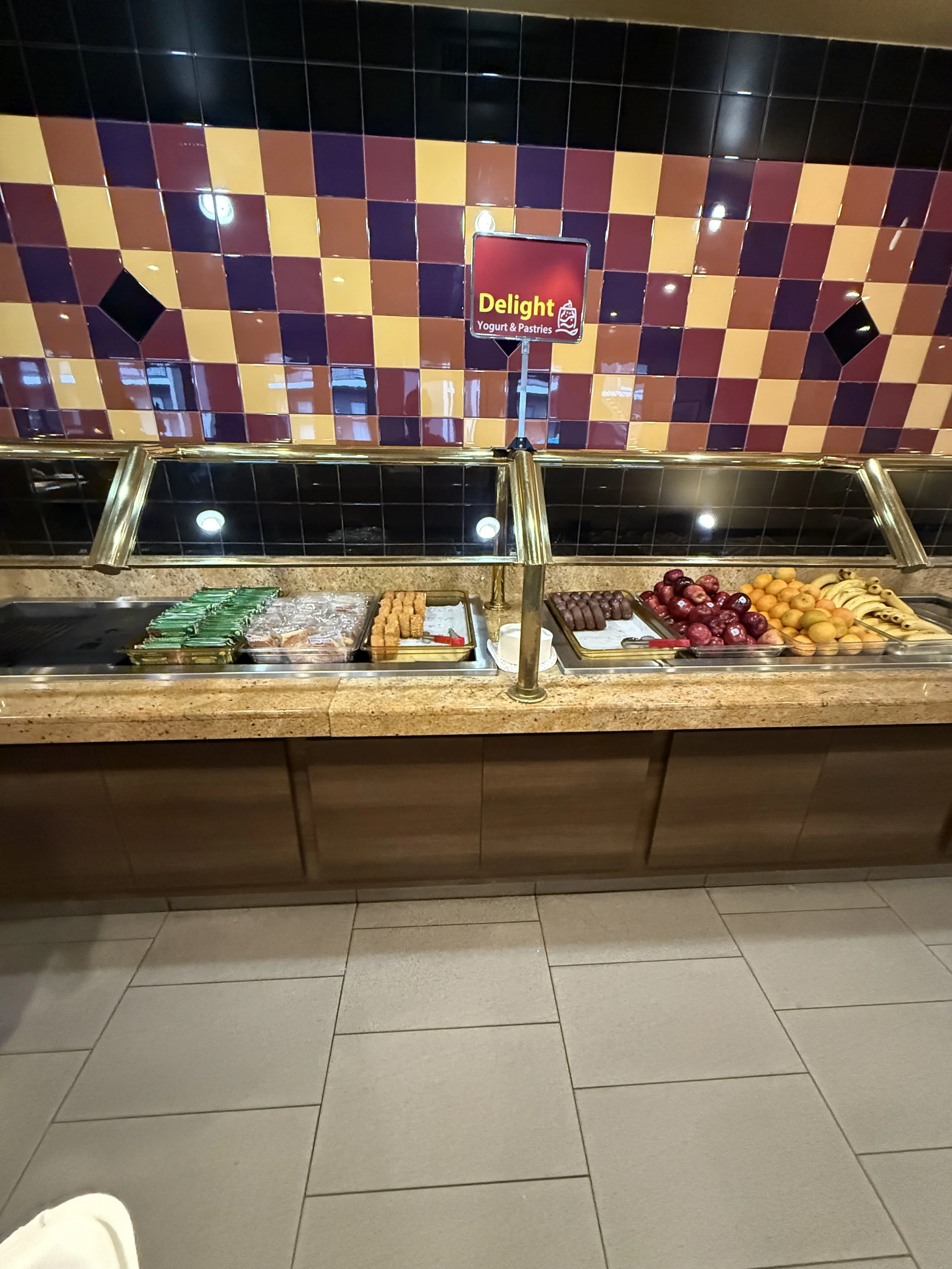 Buffet with assorted yogurt and pastries, including green wrapped items, croquettes, chocolates, red and yellow apples, and bananas, with a colorful tiled wall in the background labeled 'Delight Yogurt & Pastries'.