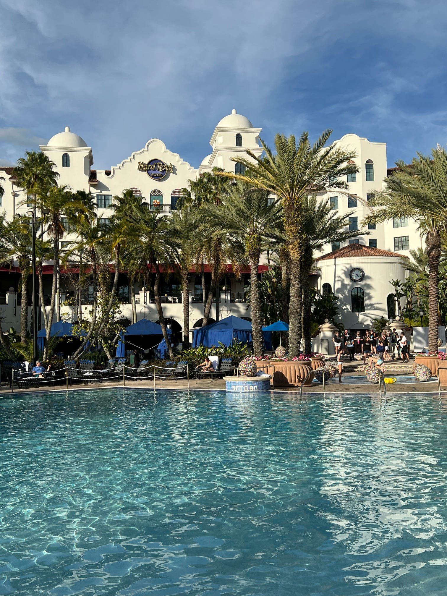 View of the Hard Rock Hotel with a pool in the foreground, surrounded by palm trees, blue umbrellas, and people relaxing and socializing outdoors.