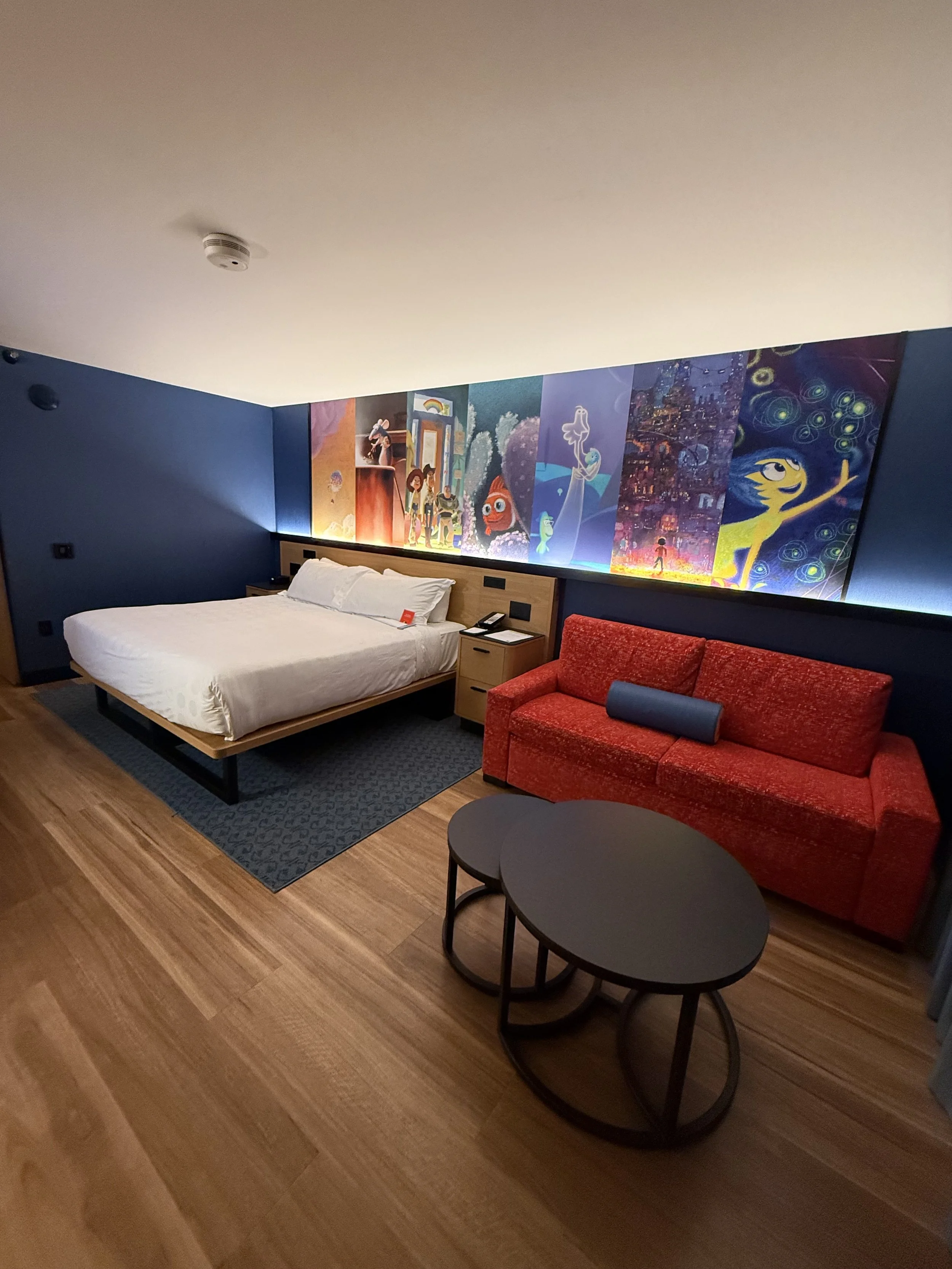 Hotel room with a large bed, red sofa, black coffee tables, wall art featuring animated characters, and a blue wall.
