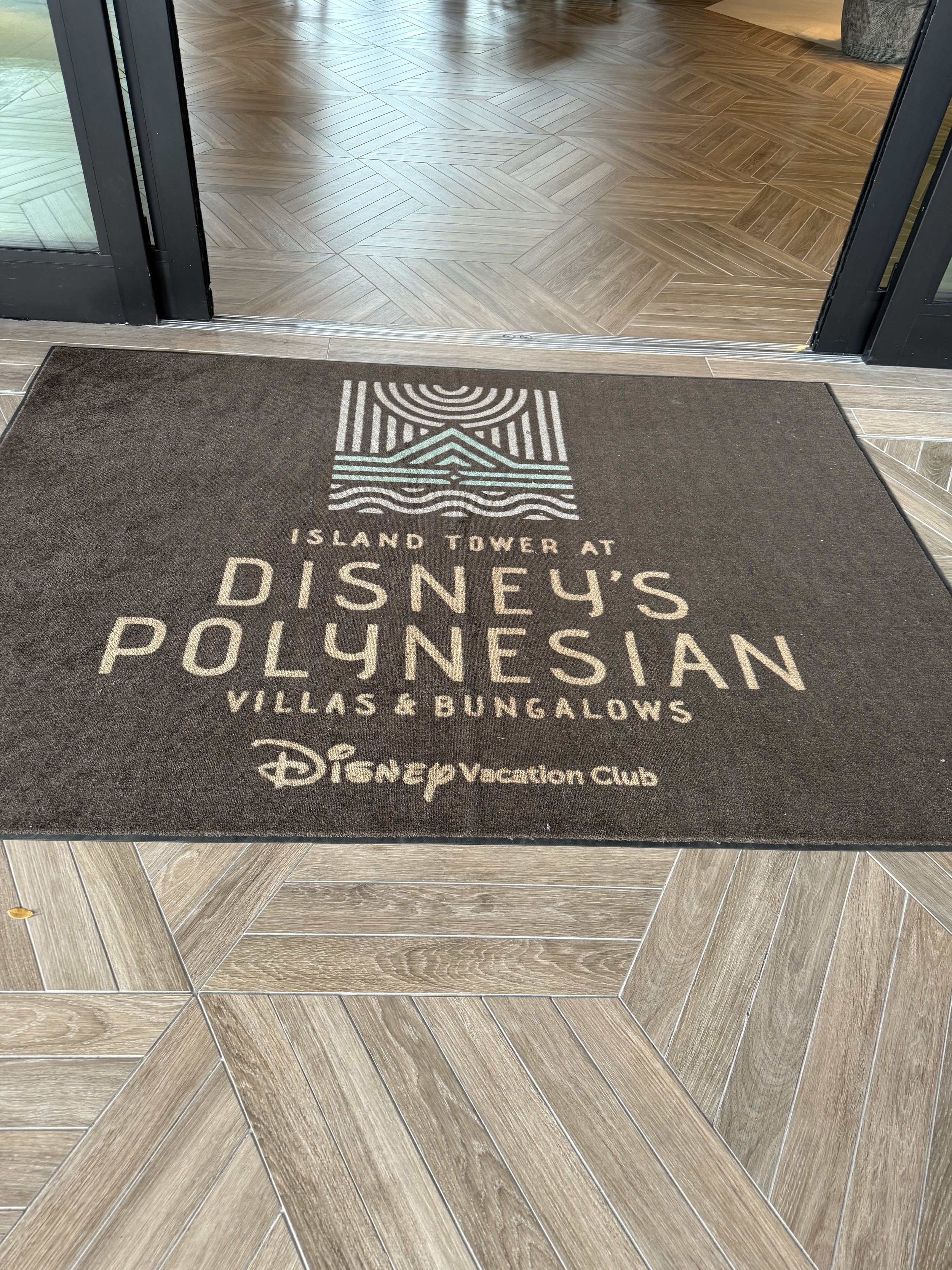 Welcome mat with text about Disney's Polynesian Villas & Bungalows at Disney Vacation Club, featuring a logo with lines and waves design, in front of a doorway with wood-patterned flooring.