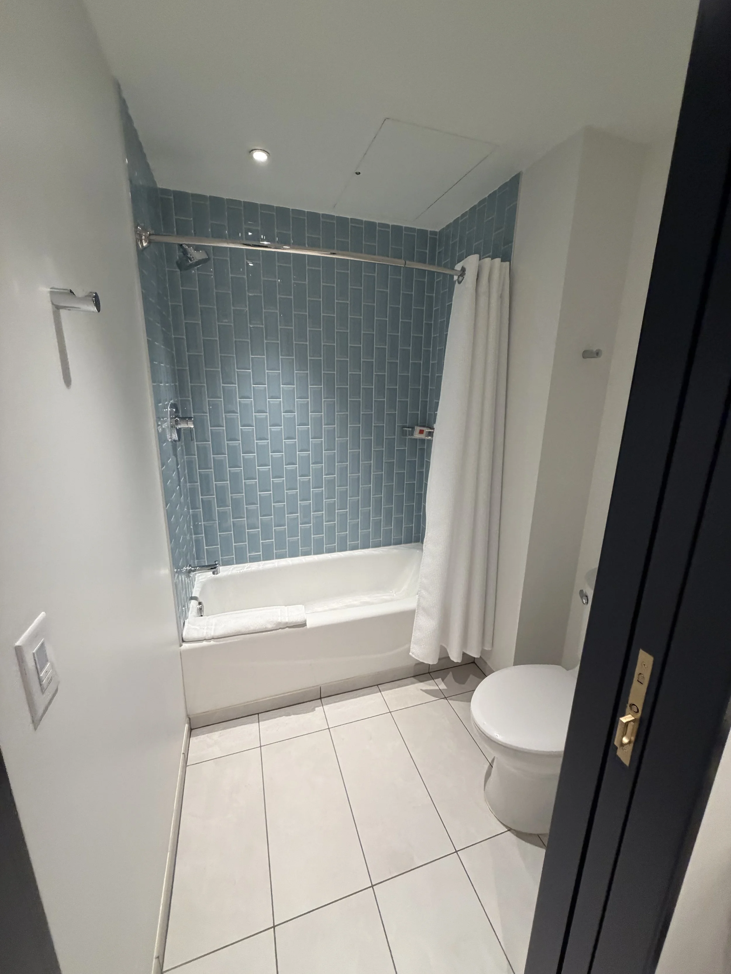 A bathroom with a bathtub and shower, blue subway tile walls, white tiles on the floor, a white toilet, and a white shower curtain.
