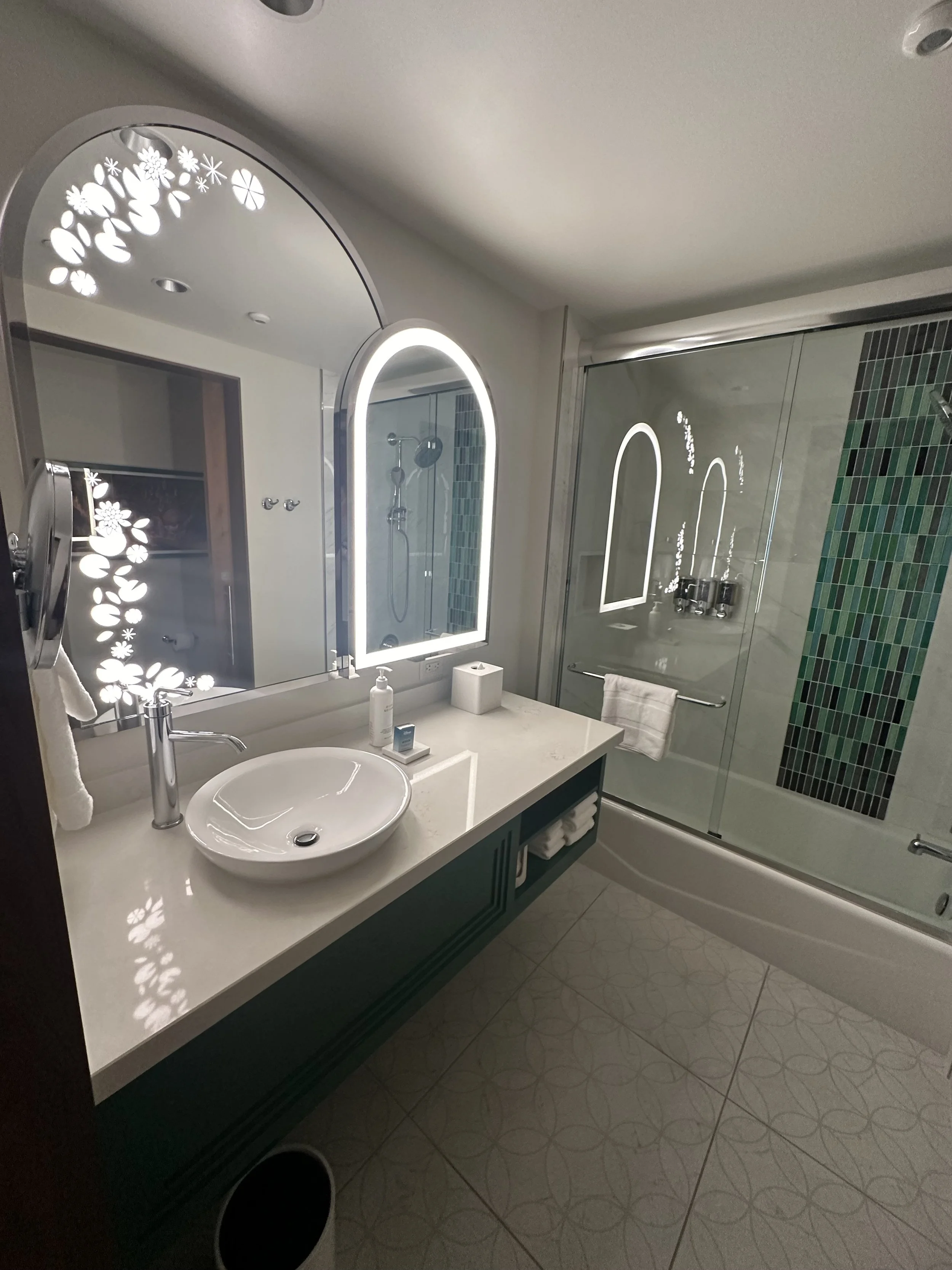 Modern bathroom with oval illuminated mirror, white sink, decorative backsplash, glass shower with mirror, and patterned floor tiles.