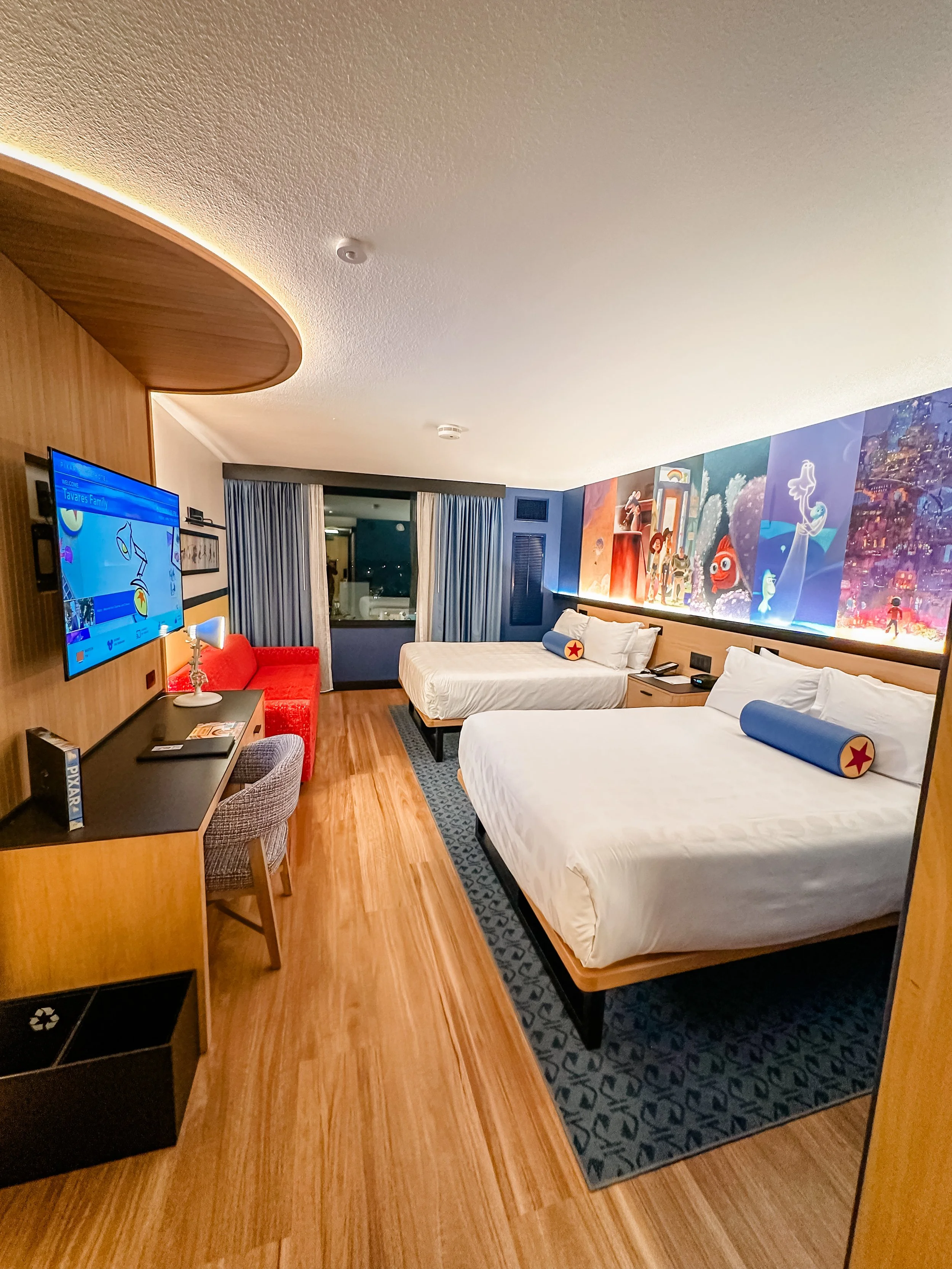 A hotel room with two beds, a large colorful mural on the wall, a television, a red armchair, a desk, and a window with curtains.