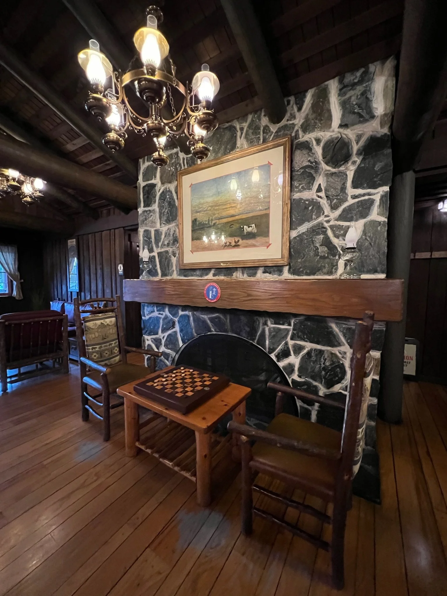A cozy room with wooden floors and walls featuring a large stone fireplace. Two vintage wooden chairs with cushioned seats sit on either side of a small wooden table with a chessboard on it. A framed landscape painting hangs above the fireplace, and 