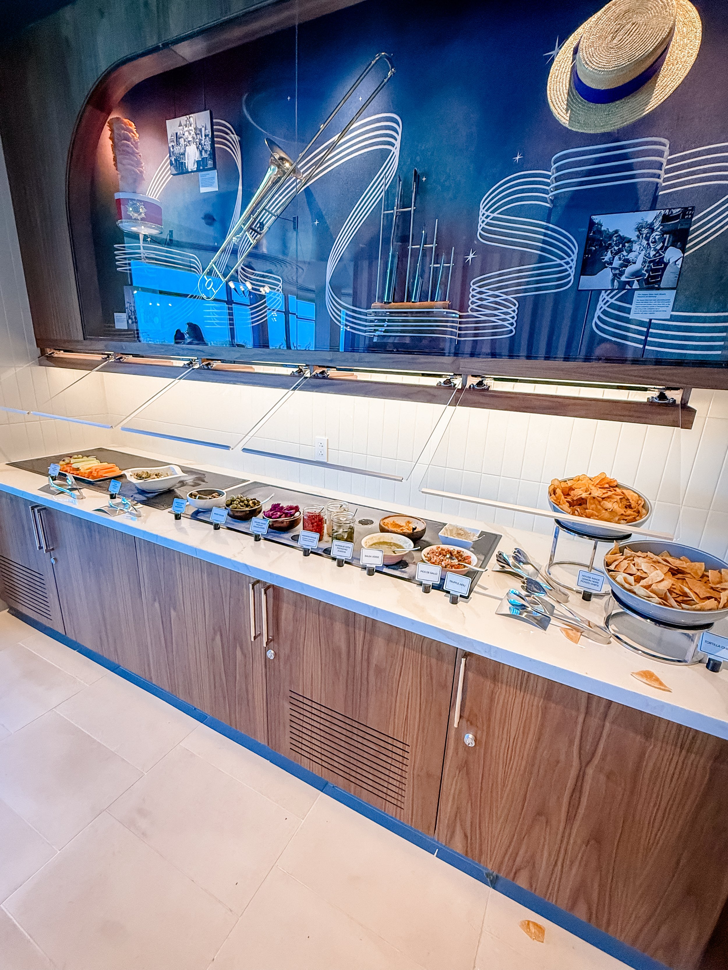 Buffet table with various Mexican food dishes like chips, salsas, and dips, with a mural of utensils and a straw hat on the wall behind.