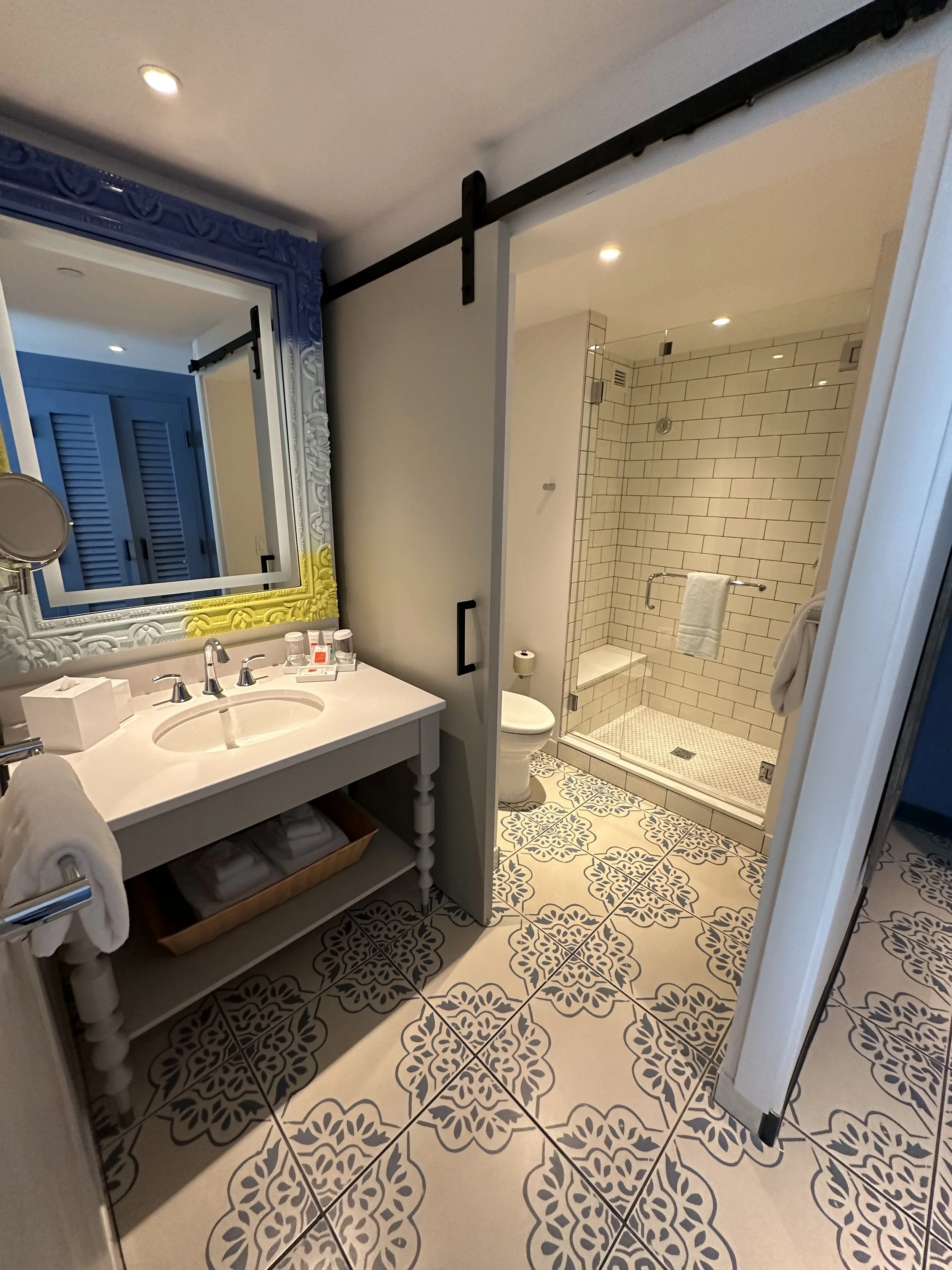 Bathroom with patterned floor tiles, a white vanity with a mirror, a shower with white tiles and a bench, a toilet, and a sliding barn door.