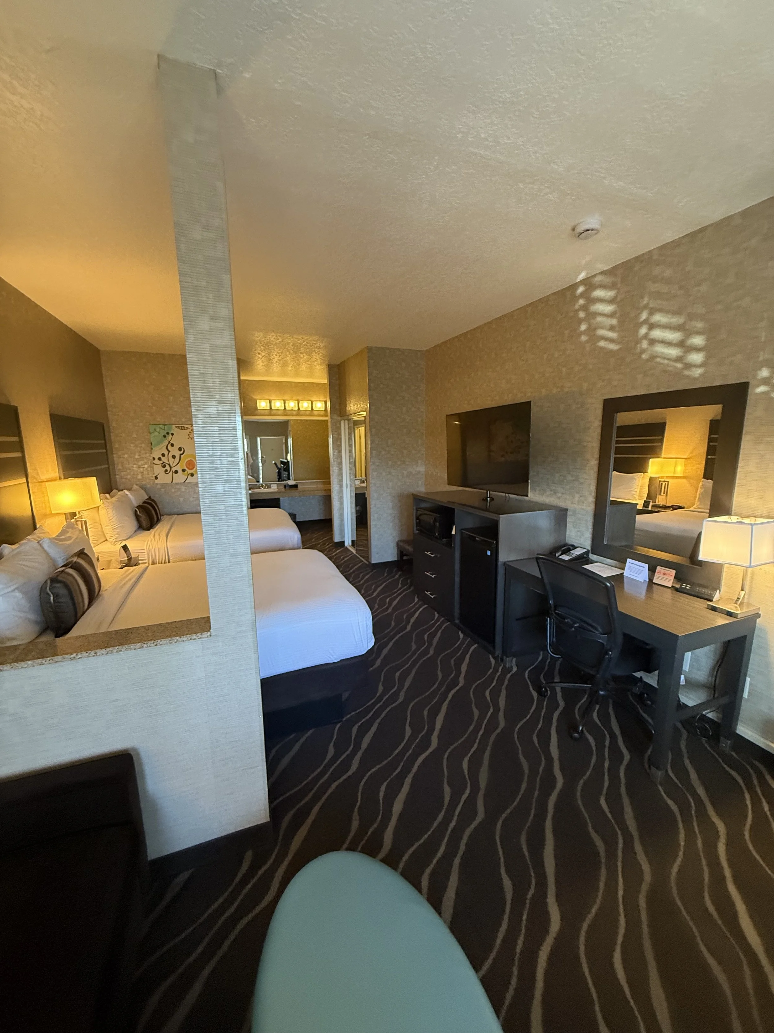 A hotel room with two beds, a desk with a lamp and mirror, a TV, and a bathroom in the background. The room has patterned carpet and neutral-colored walls.