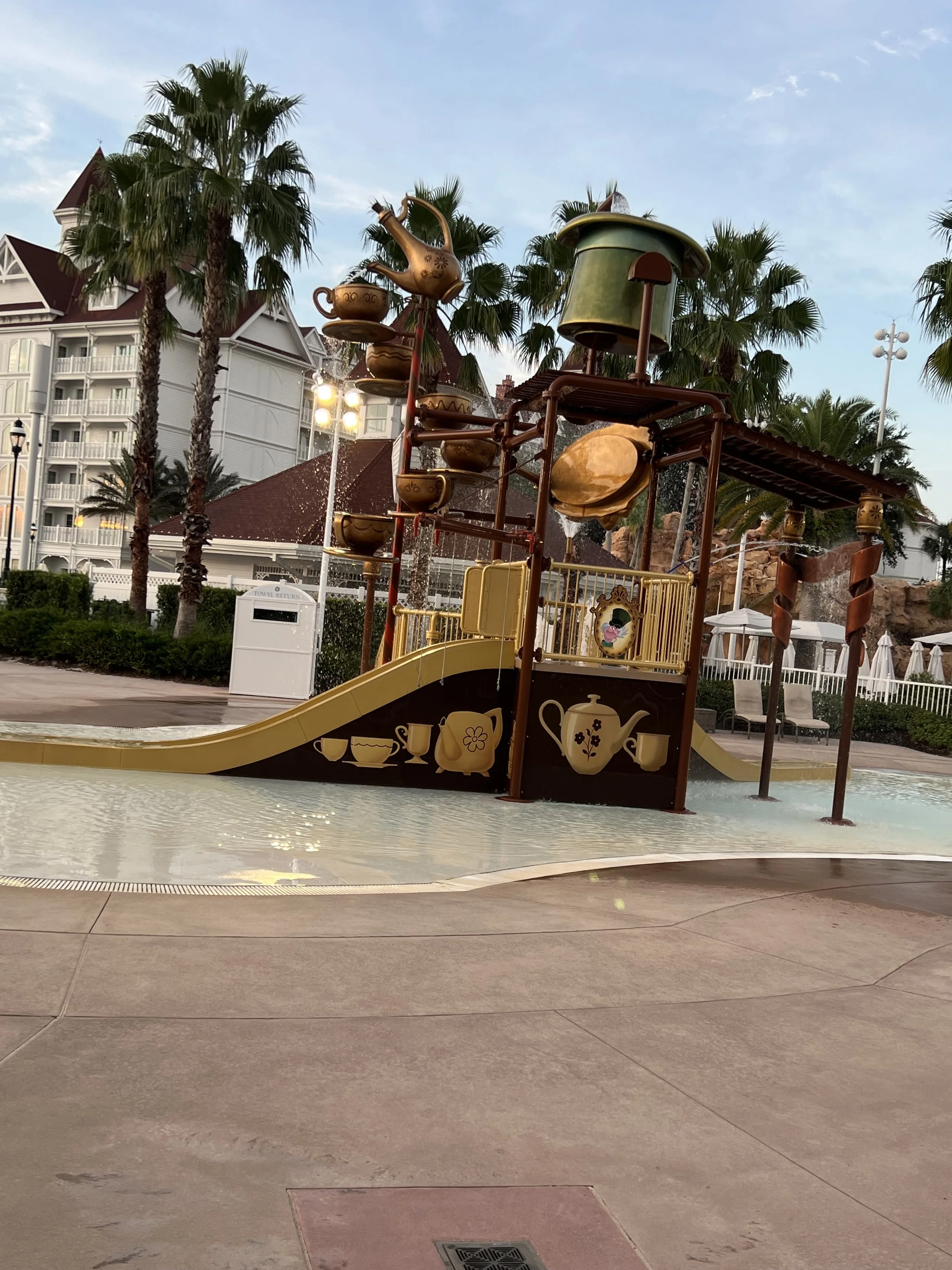 A children's water playground with a tea party theme, featuring a slide, teapot, cups, and teacup-shaped water features, surrounded by palm trees and a resort backdrop.