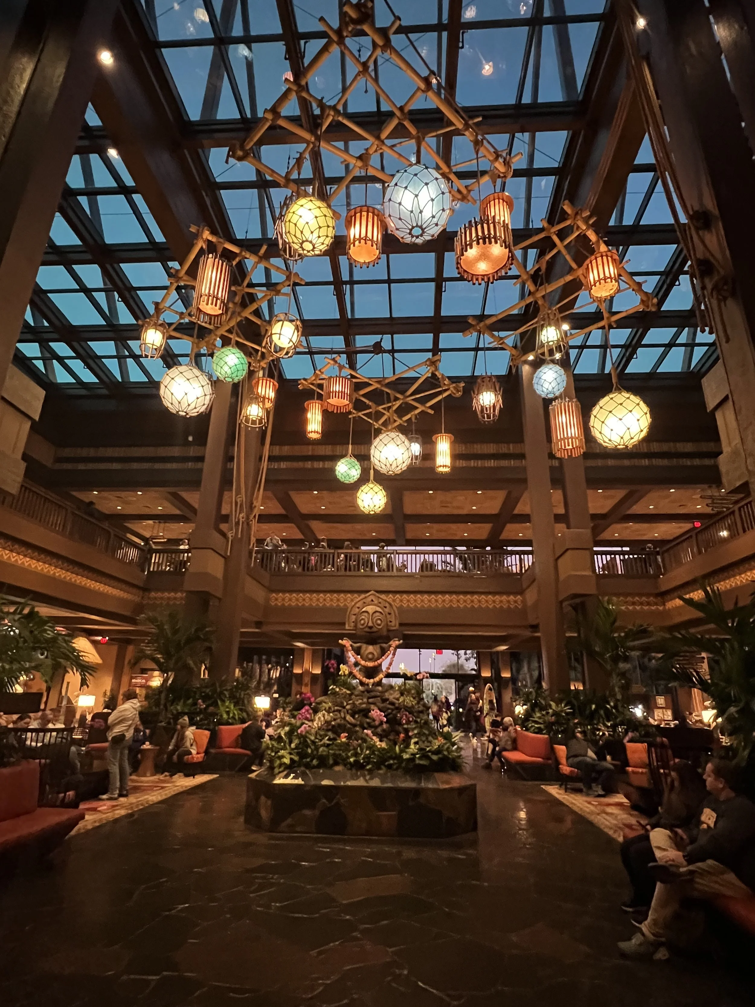 Interior of a spacious hotel lobby with a high glass ceiling, hanging lantern-style lights, a central decorative fountain with tropical plants and flowers, and seating areas with orange and patterned cushions, filled with guests and visitors.