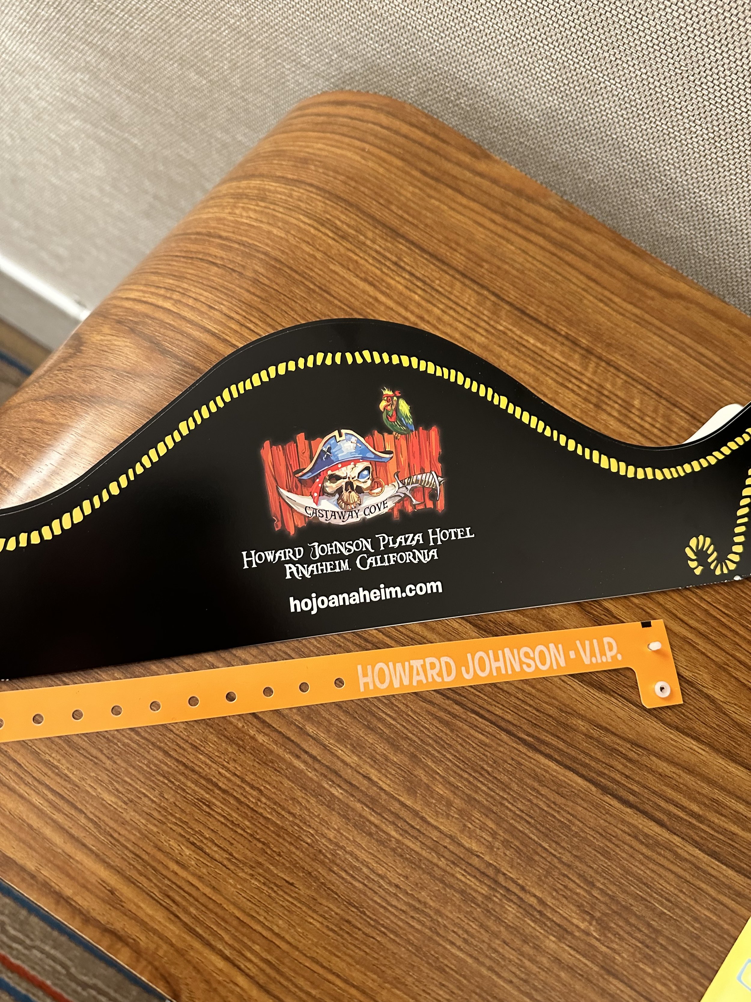 A black plastic coaster with pirate-themed graphics and text, placed on a wooden surface. Also, a yellow wristband with the inscription 'HOWARD JOHNSON - V.I.P.' is beside it.