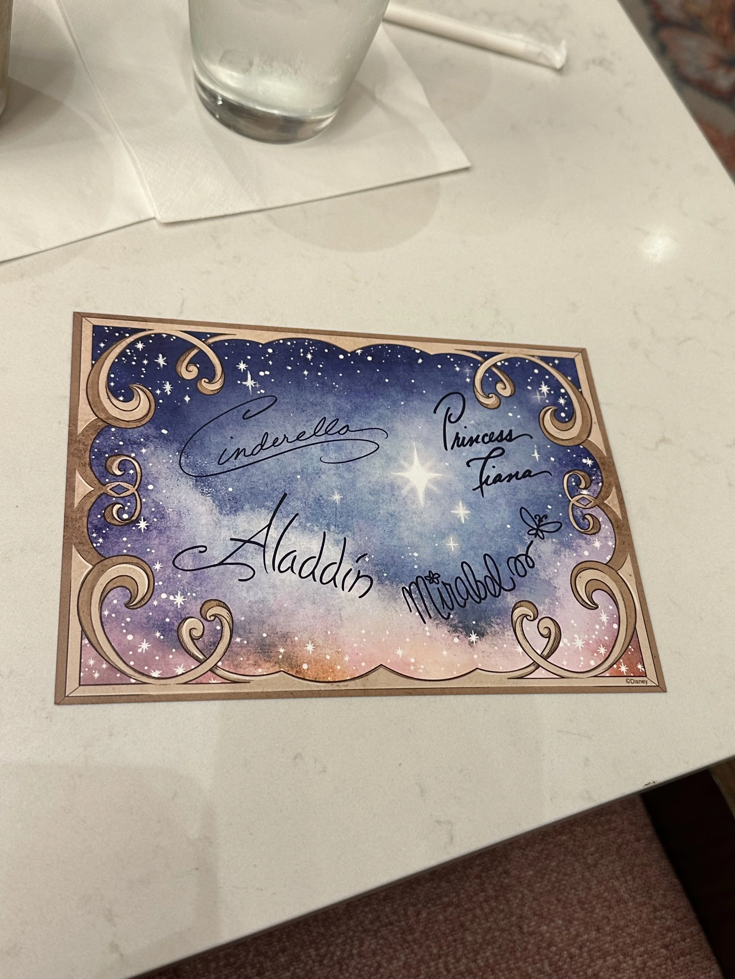 A colorful paper with a starry sky background and golden decorative borders, featuring handwritten signatures of Disney princesses including Cinderella, Princess Fiona, Aladdin, and Mirabel.