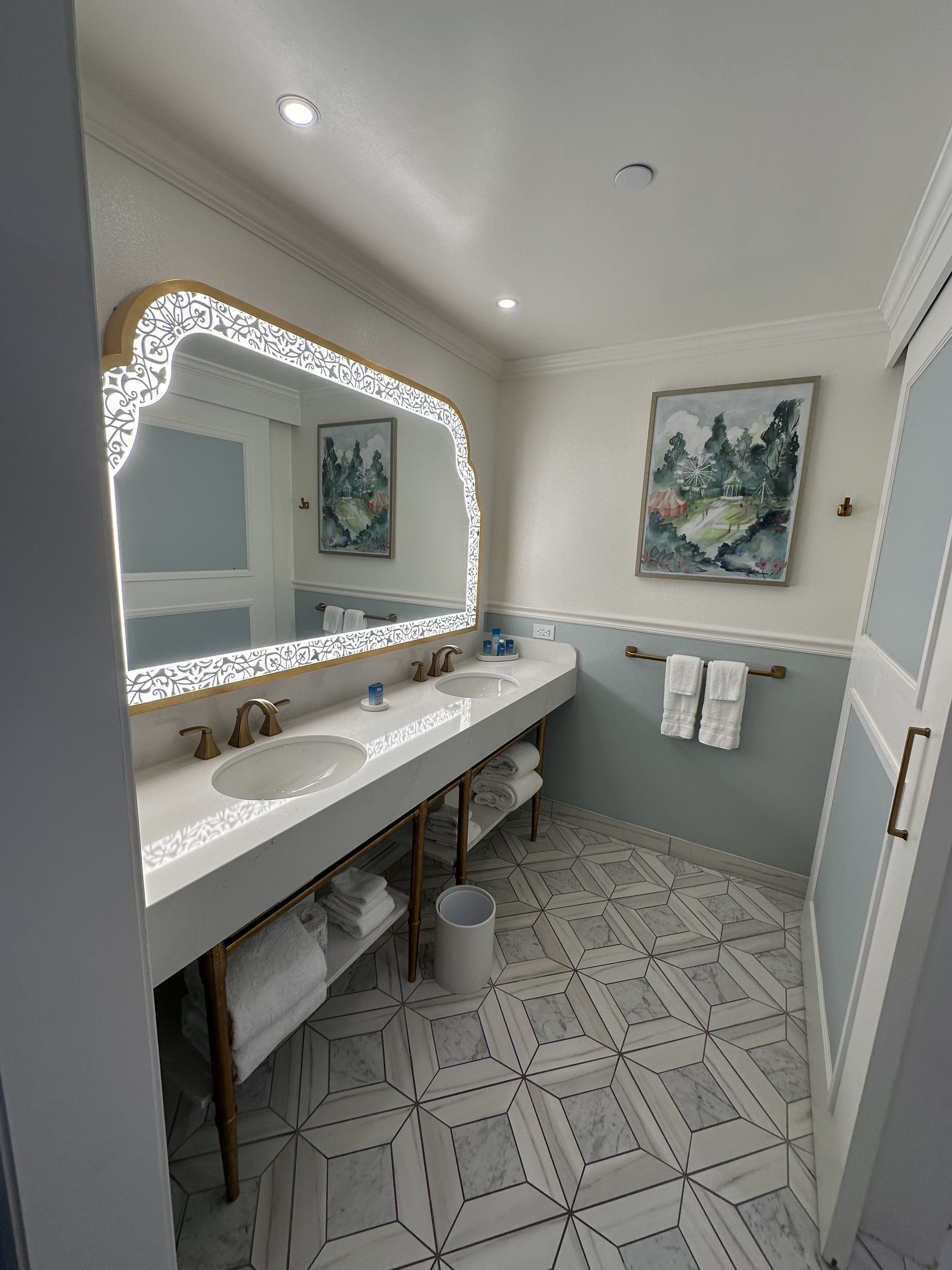 A modern bathroom featuring a double sink vanity with a large mirror framed with decorative lighting, a painting on the wall, and neatly folded towels on shelves beneath the countertop.