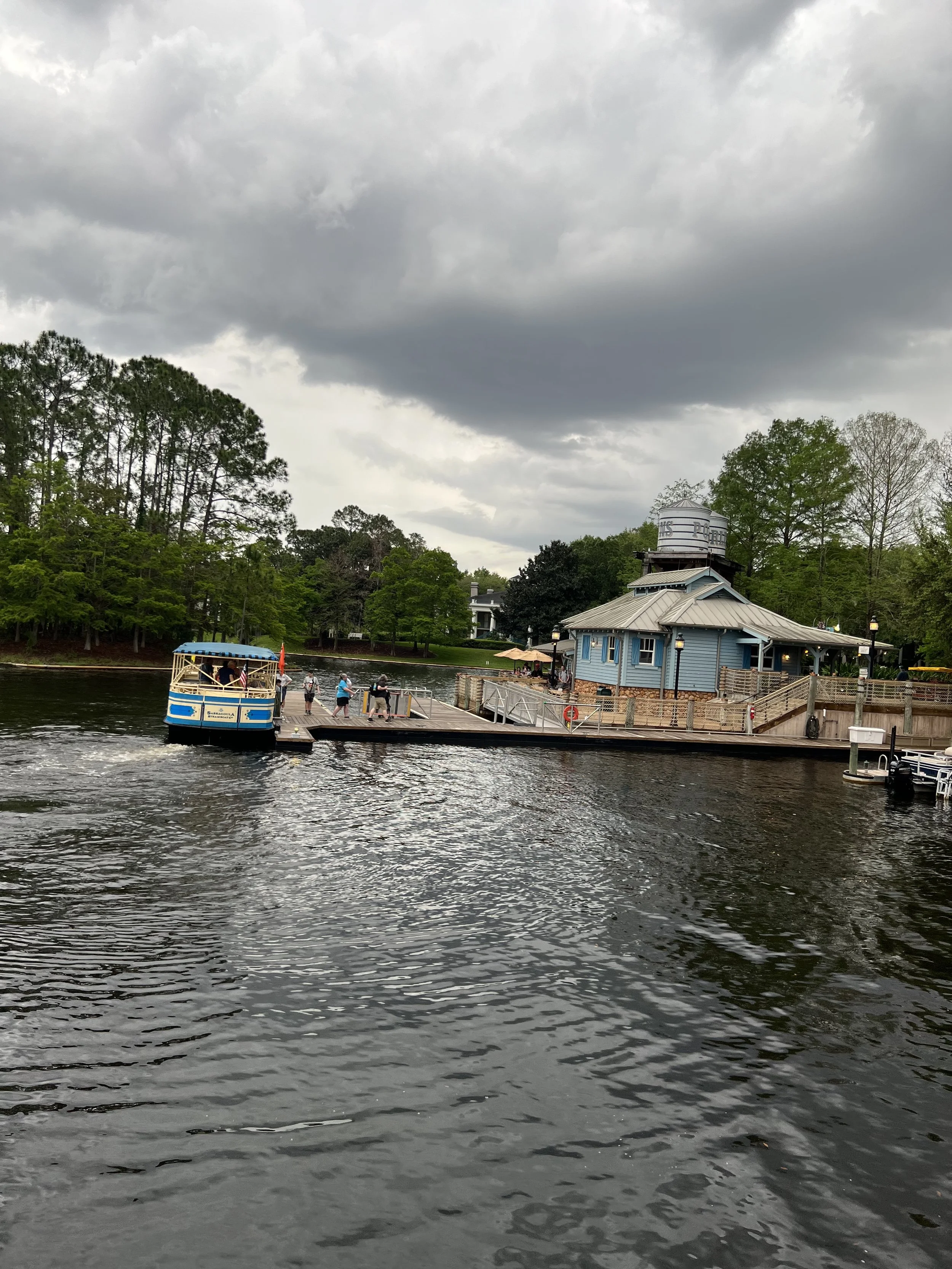 A dock on a river with several people boarding a small boat, next to a blue building with a water tower on top, surrounded by trees under a cloudy sky.