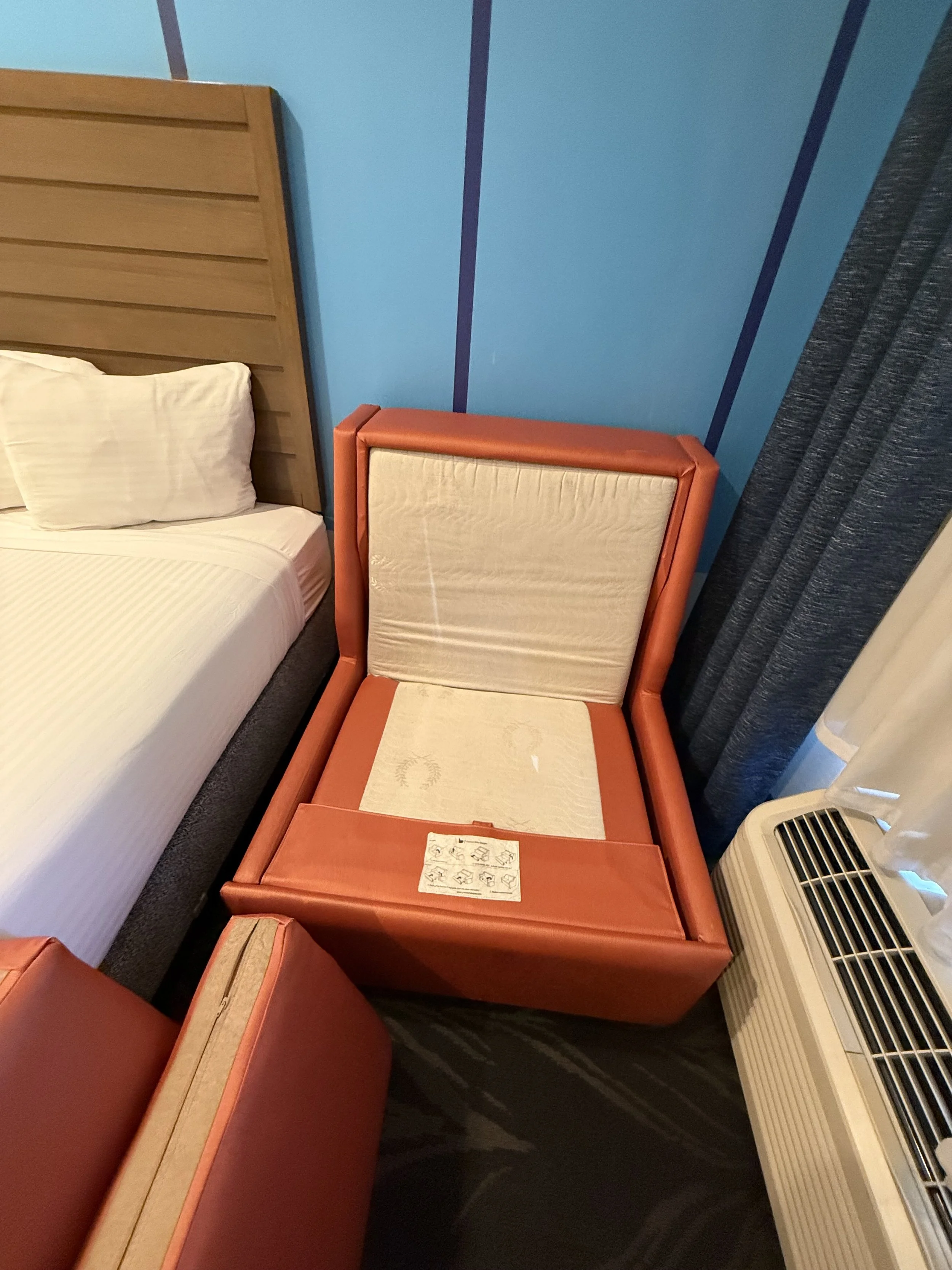 Unopened orange bed with floral patterned mattress inside a hotel room, next to a white pillow, with a wooden headboard, blue wall, dark curtains, and an air conditioning unit.