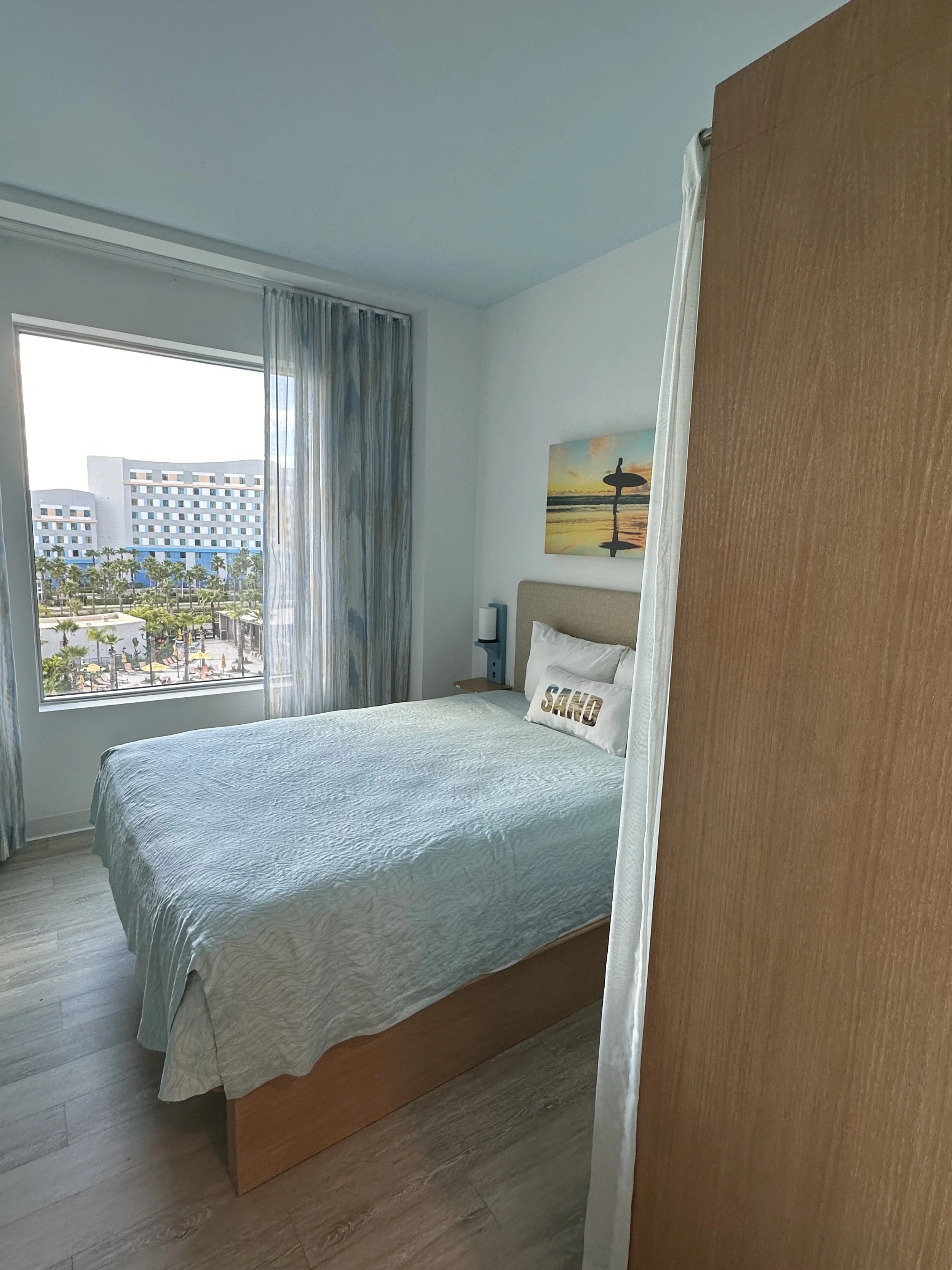 Tourist hotel room with a bed, nightstand, art of a surfer at sunset above the headboard, window with view of buildings and palm trees, and partial view of a wooden closet door.